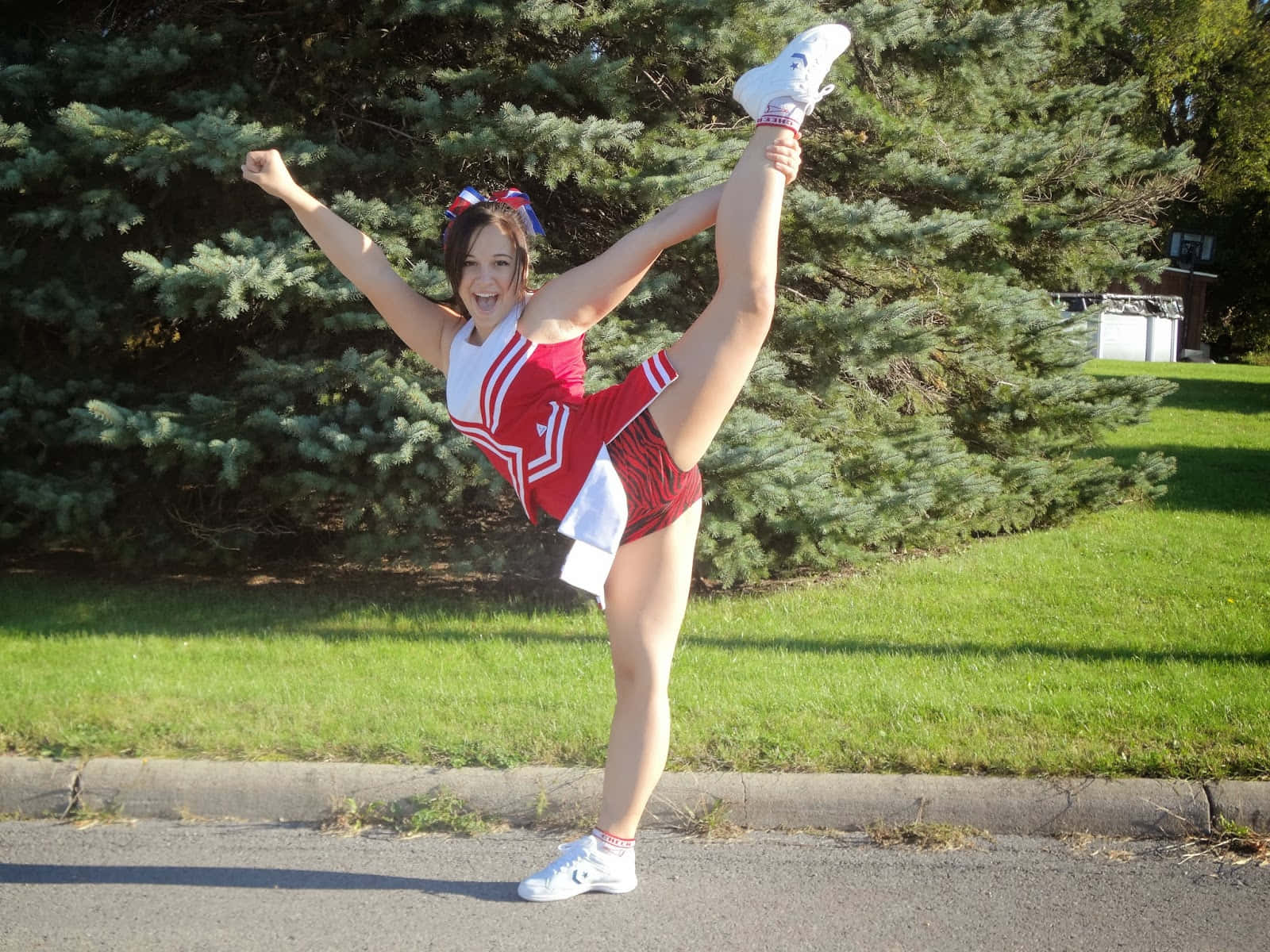 Cheer 桌布 壁紙 — 一位微笑的啦啦隊員身穿紅白制服，在陽光明媚的街道上表演高踢腿，身後有綠樹。