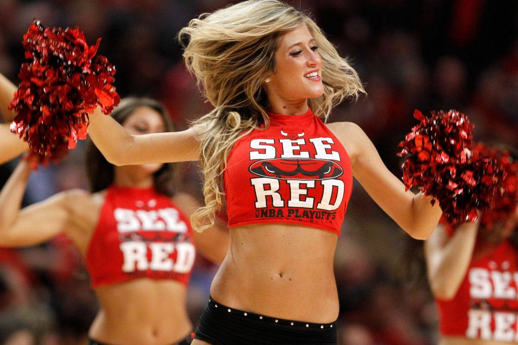 Cheerleader 桌布 壁紙 — 一位微笑的啦啦隊員身穿紅色短上衣和黑色褲子，在籃球比賽中手持紅色絨球跳舞。