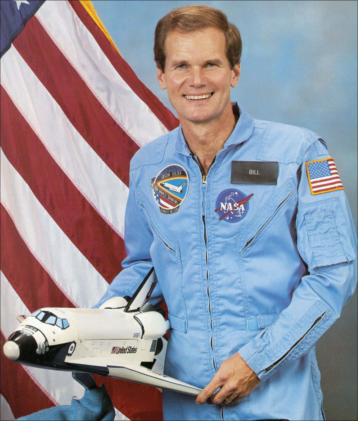 NASA 動態圖片 桌布 壁紙 — 一位微笑的太空人穿著藍色NASA飛行服，在美國國旗前拿著太空梭模型。