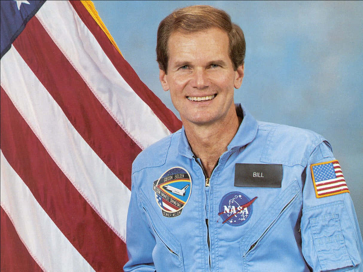 NASA 動態圖片 桌布 壁紙 — 一位微笑的太空人身穿藍色NASA飛行服，站在美國國旗前，背景是柔和的藍色。