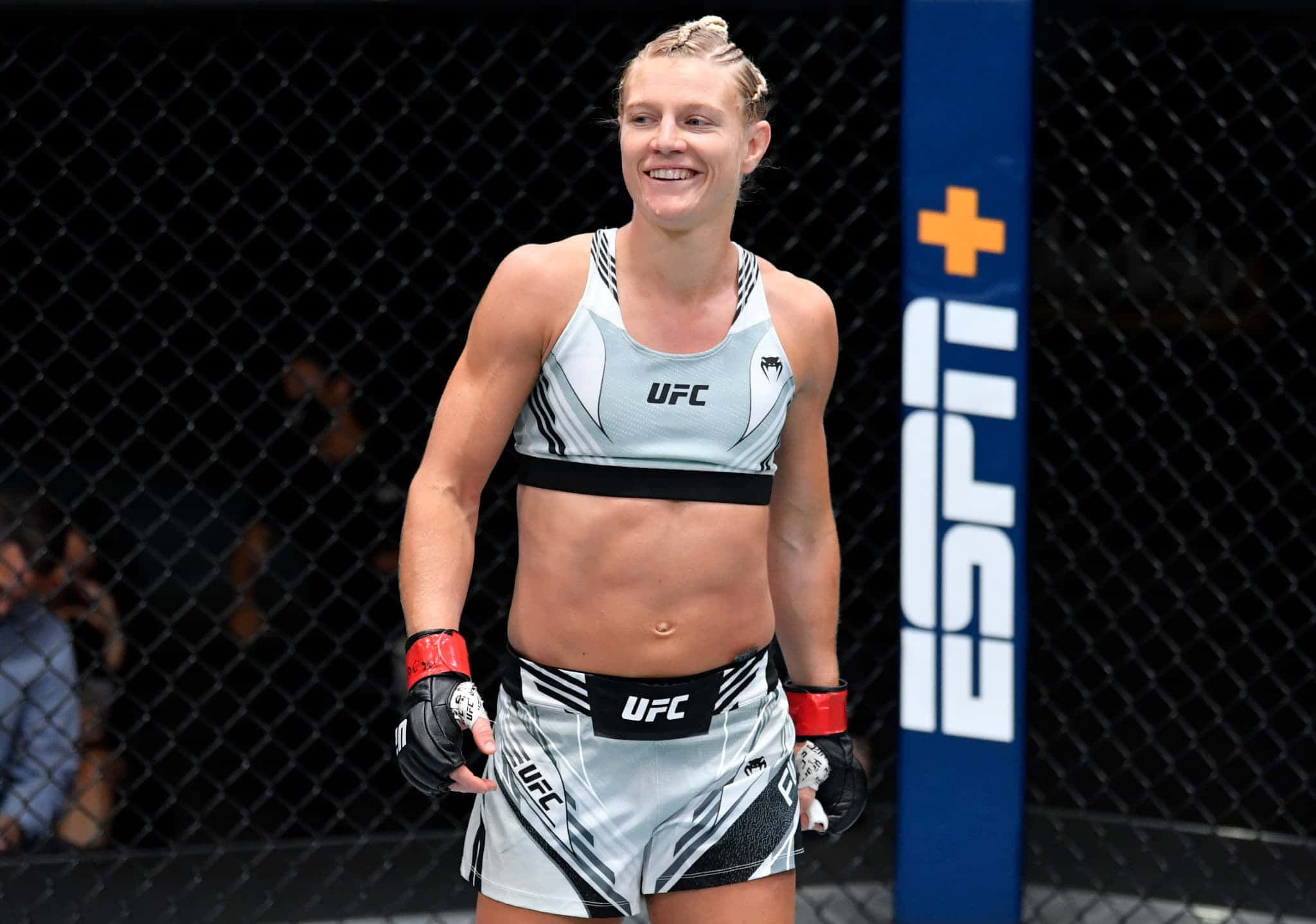 桌布 壁紙 — 一位微笑的女MMA拳手穿著UFC裝備，自信地站在八角籠內，ESPN+品牌標誌清晰可見。