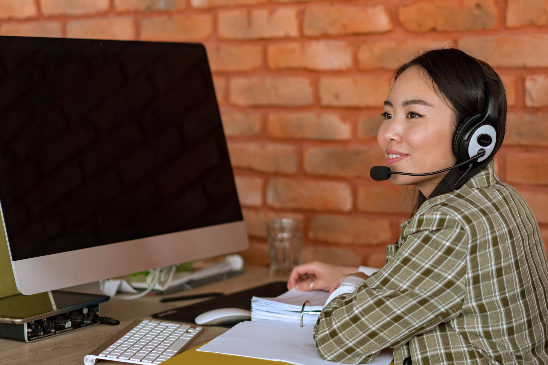 Working 桌布 壁紙 — 一位微笑的女人穿著格子襯衫，戴著耳機，在電腦和磚牆背景的辦公桌前工作。