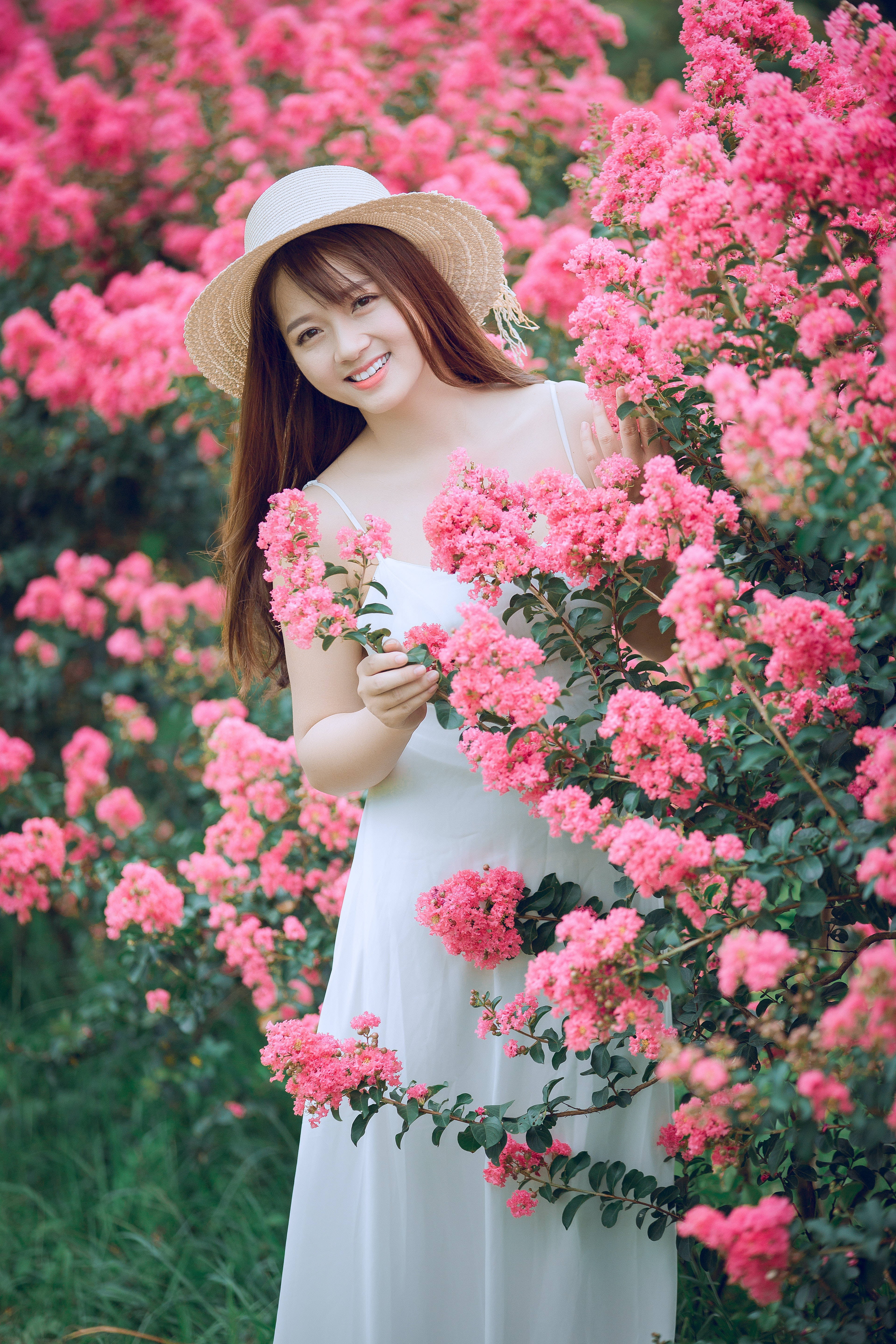 girls 桌布 壁紙 — 一位微笑的女人穿著白色連衣裙和草帽，站在一片充滿活力的粉紅色開花灌木叢中。