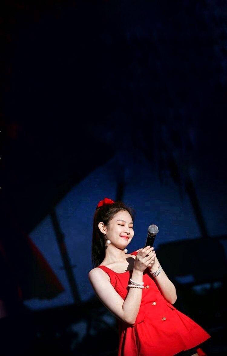 Jennie 桌布 壁紙 — 一位微笑的女人穿著紅色連衣裙，在舞台燈光下拿著麥克風，背景黑暗。