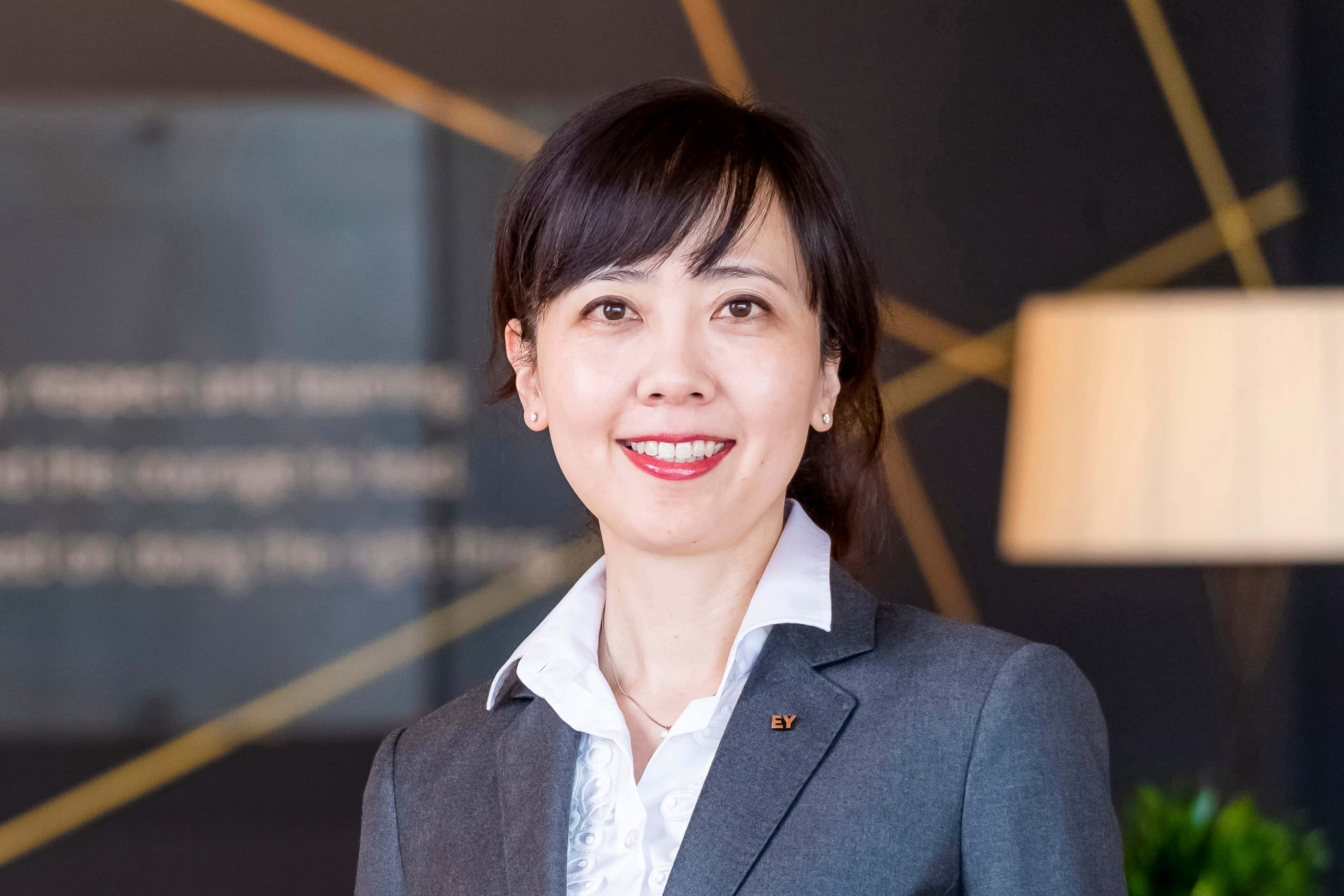UFC 桌布 壁紙 — 一位微笑的女商人穿著灰色西裝和白色襯衫，自信地站在一個現代辦公室，帶有幾何金色牆壁裝飾。