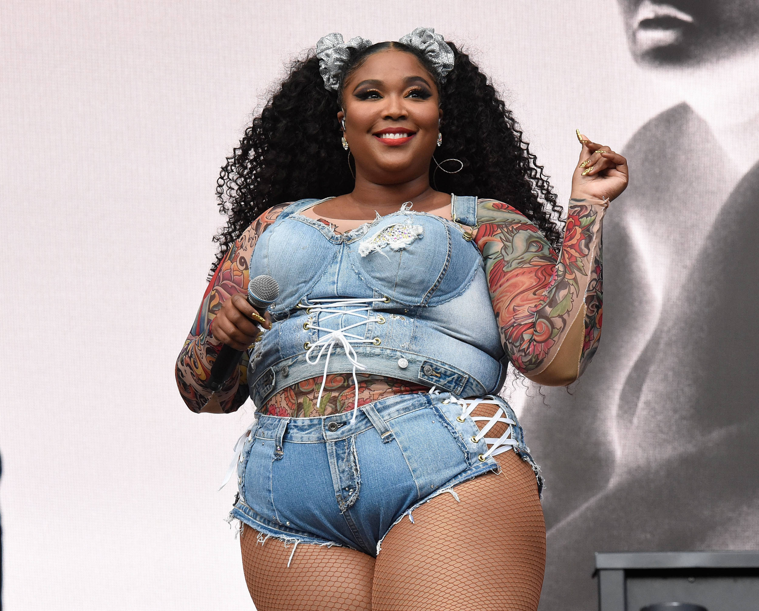 Lizzo 桌布 壁紙 — 一位微笑的女士，擁有捲曲頭髮和色彩繽紛的刺青，穿著牛仔服裝和漁網襪在舞台上表演。
