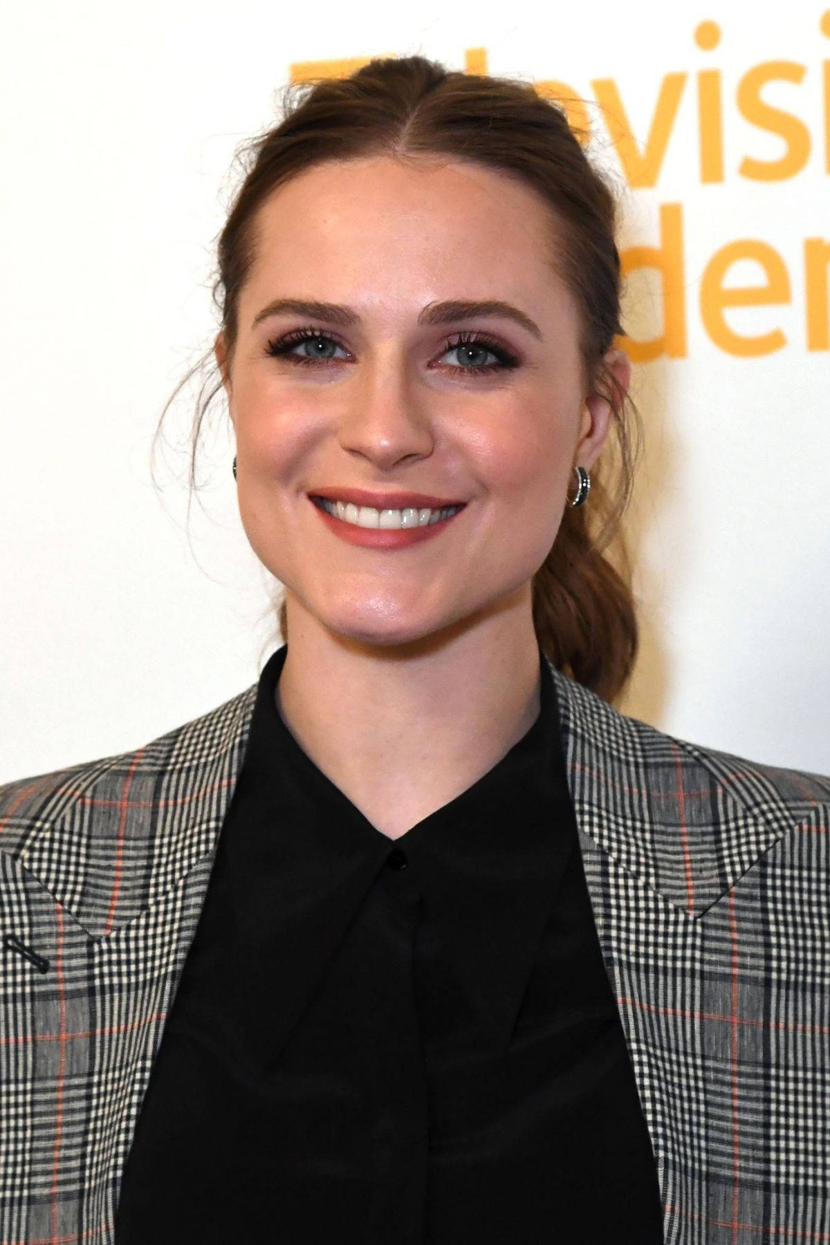 Celebrity 桌布 壁紙 — 一位微笑的女士，棕色頭髮紮成馬尾，在電視活動中穿著格紋西裝外套，內搭黑色襯衫。