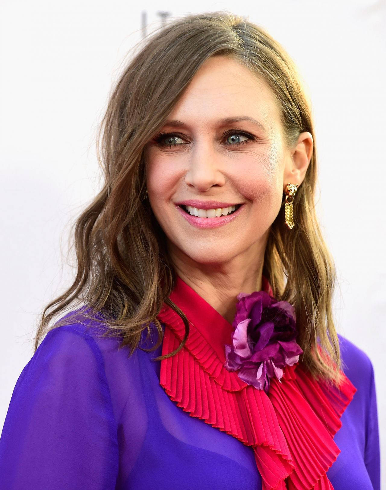 Celebrity 桌布 壁紙 — 一位微笑的女士，波浪形棕色頭髮，穿著充滿活力的紫色襯衫，搭配大膽的紅色褶邊領和紫色花朵飾品。