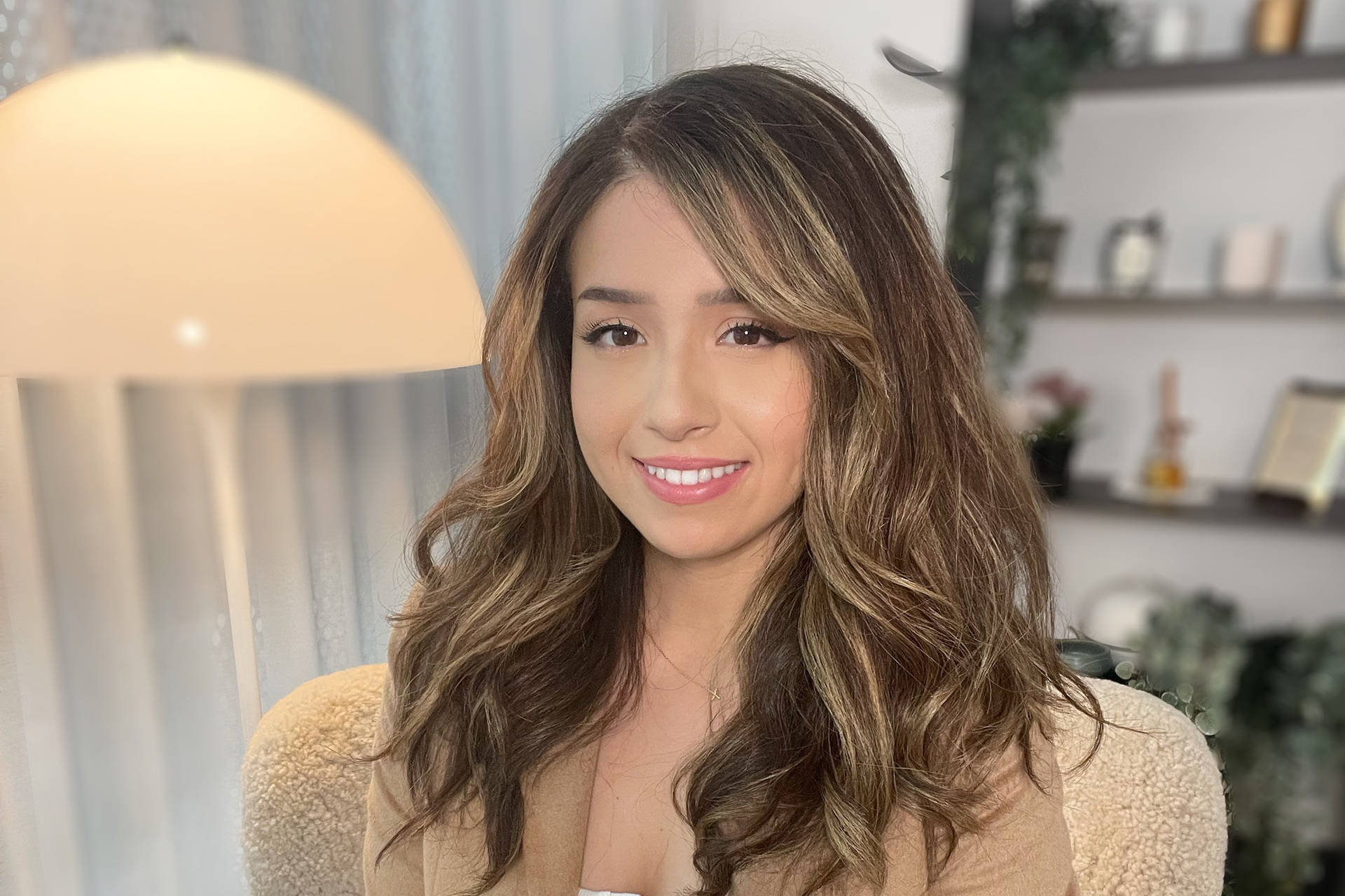 pokimane 桌布 壁紙 — 一位微笑的女士，波浪棕色頭髮，坐在一個由溫暖圓頂燈照亮的舒適房間裡。