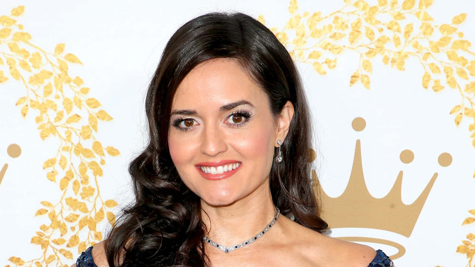 Danica Mckellar 桌布 壁紙 — 一位微笑的女士，深色波浪髮，佩戴優雅珠寶，站在白色背景前，背景飾有金葉圖案和皇冠。