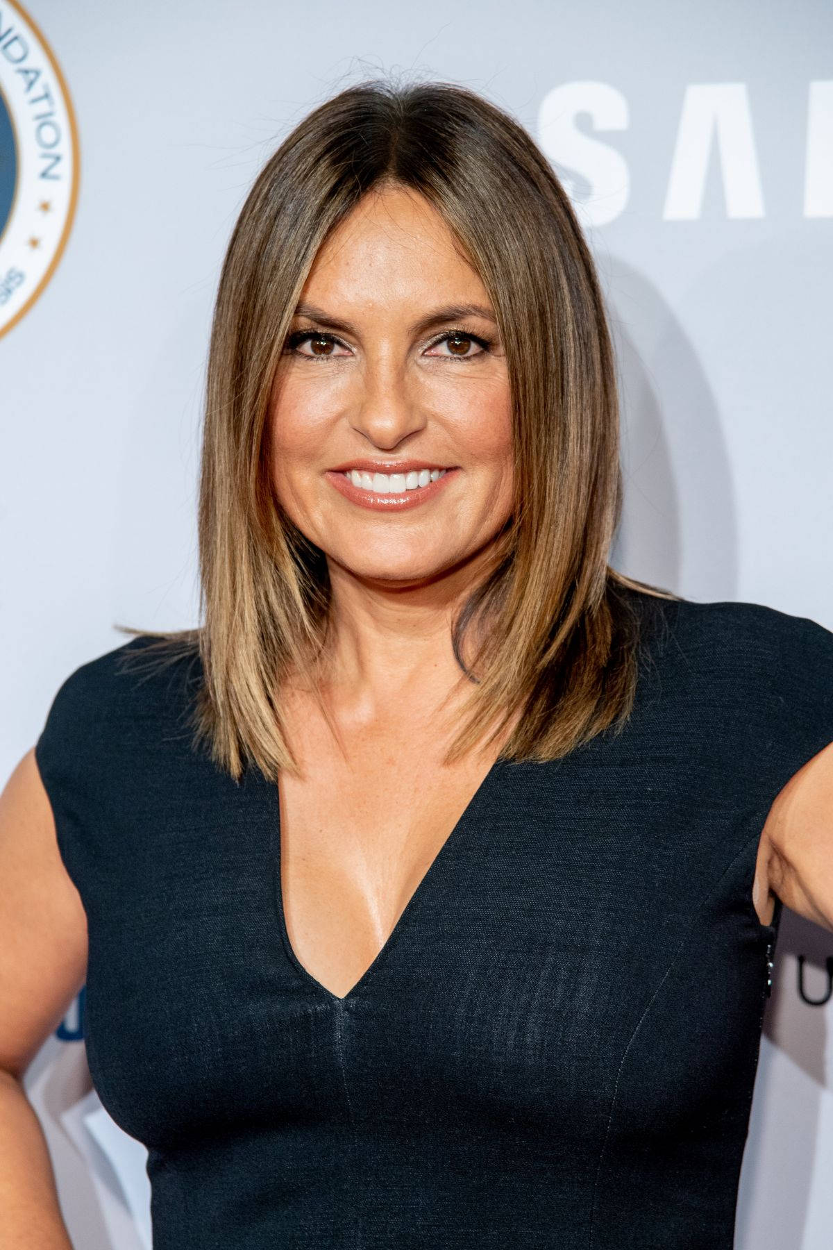 Celebrity 桌布 壁紙 — 一位微笑的女士，留著及肩棕色頭髮，在一個有品牌背景的正式活動中穿著黑色連衣裙。