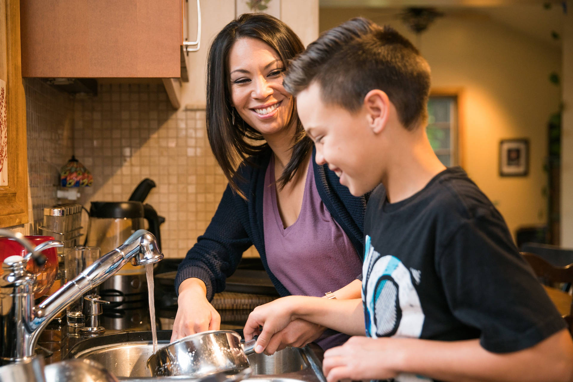 Family 桌布 壁紙 — 一位微笑的女士和男孩在溫暖燈光的廚房水槽中一起洗碗。