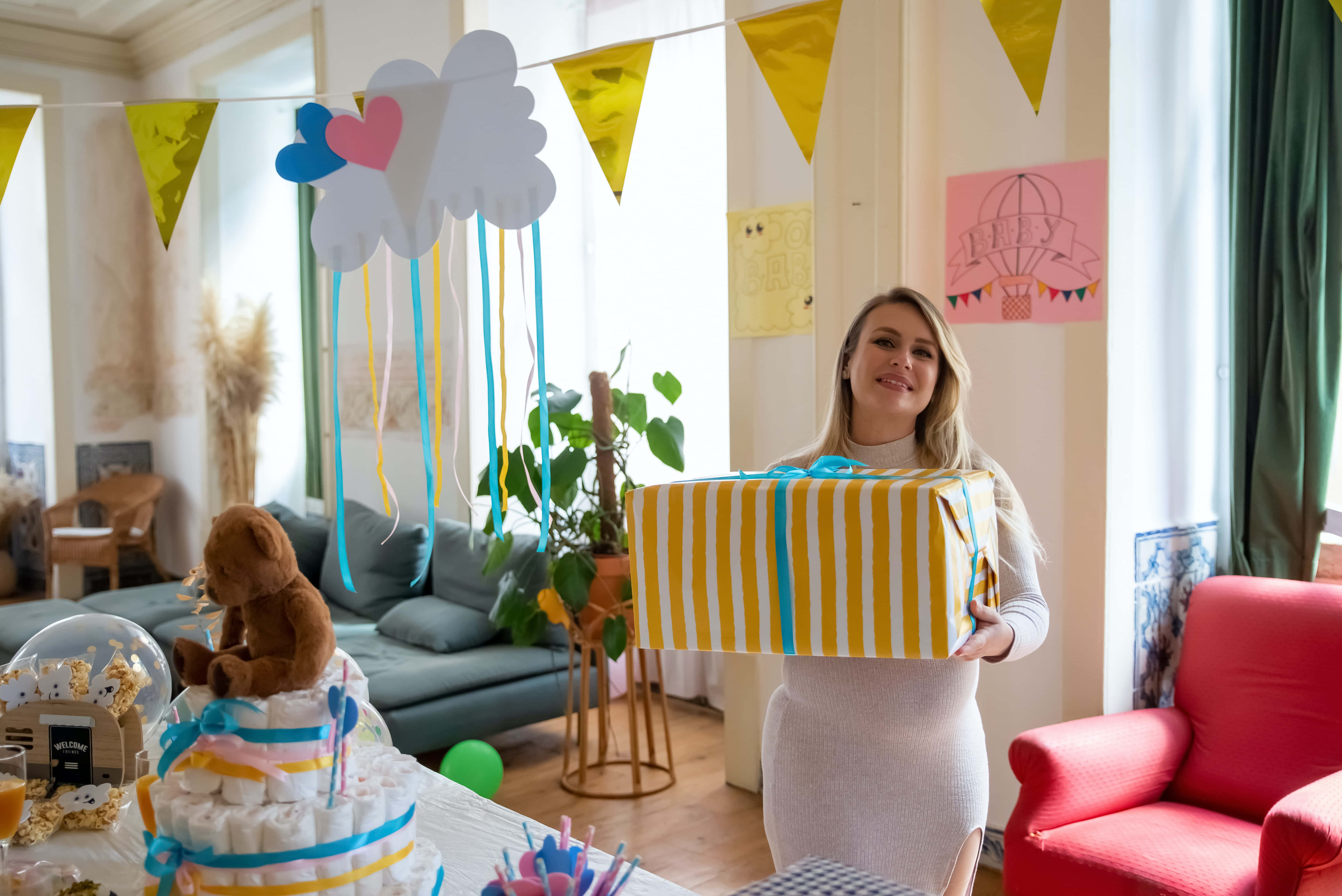 Baby Shower 桌布 壁紙 — 一位微笑的女士在一個以雲朵、愛心和彩旗裝飾的彩色嬰兒派對上，拿著一個黃色條紋禮物盒。