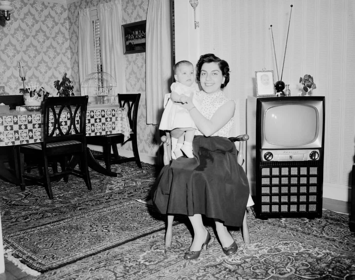 1950s 桌布 壁紙 — 一位微笑的女士在一個帶有圖案壁紙和舊電視的復古客廳中抱著嬰兒。