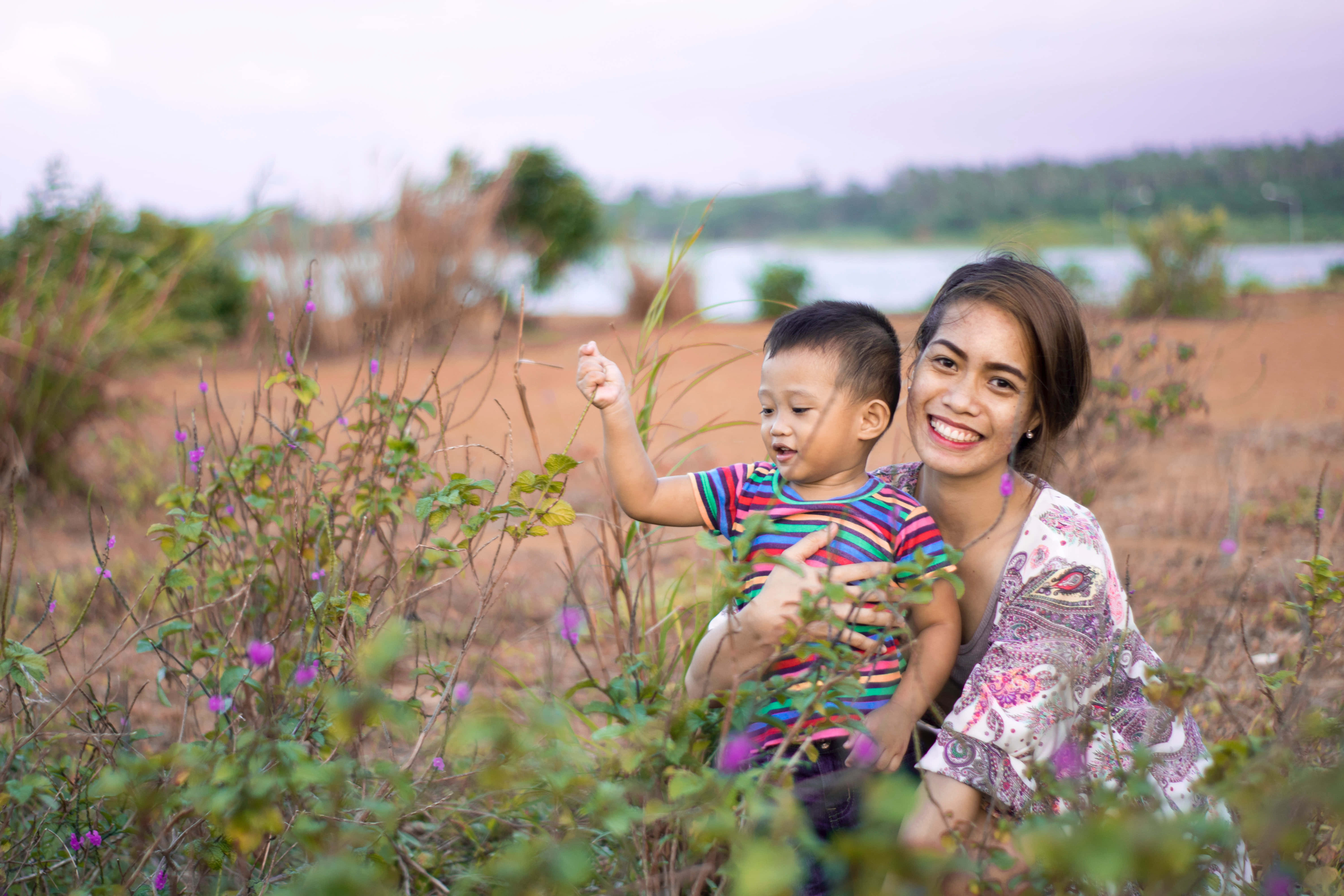 Mom 桌布 壁紙 — 一位微笑的女士在柔和的日光下，於平靜的湖邊紫色野花中抱著一個好奇的幼兒。