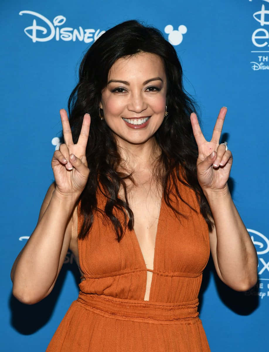 Ming Na Wen 桌布 壁紙 — 一位微笑的女士穿著橙色連衣裙，在藍色迪士尼背景下做出和平手勢。