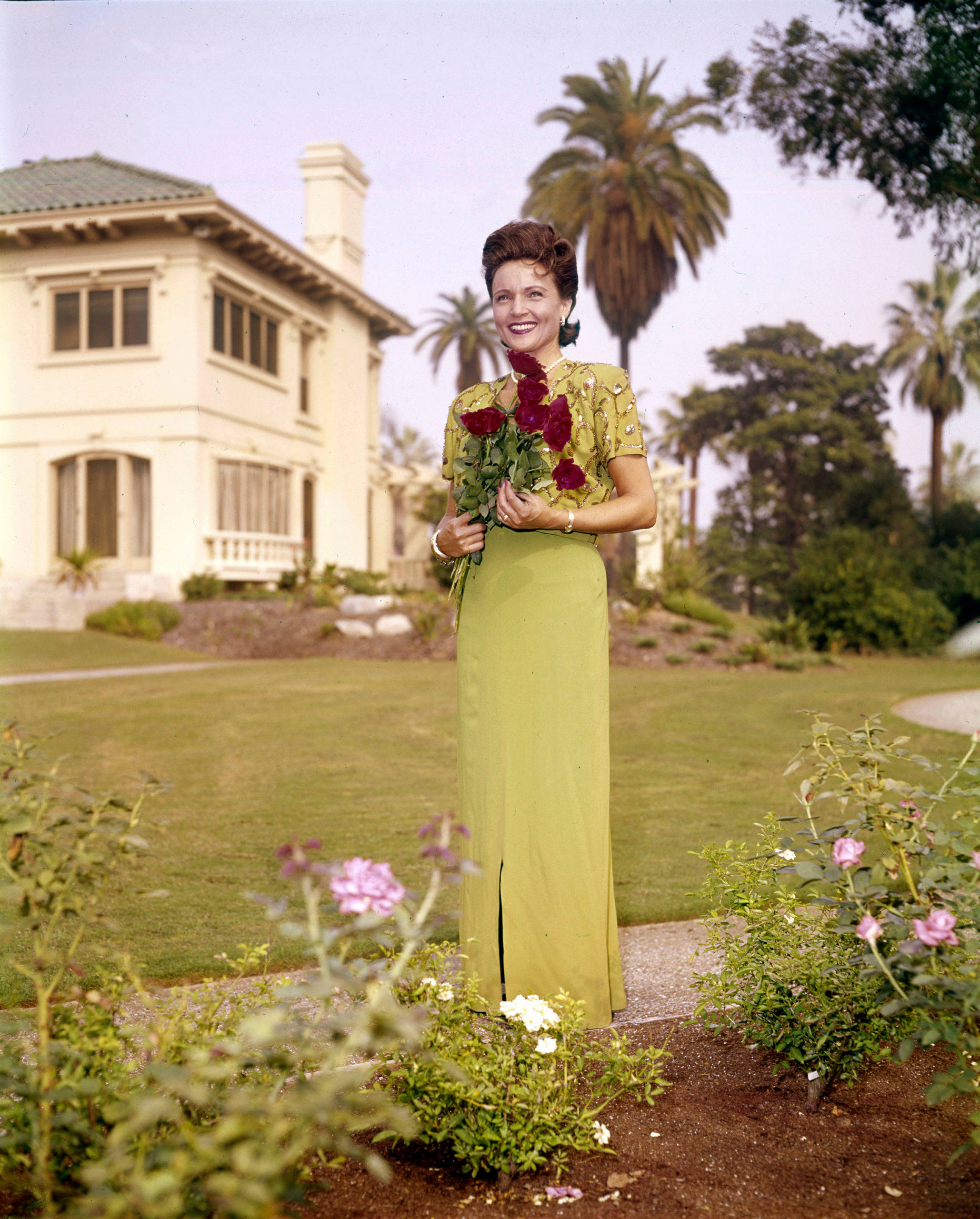 Betty White 桌布 壁紙 — 一位微笑的女士穿著綠色連衣裙，在種有棕櫚樹的白色房子前的花園裡拿著紅玫瑰。