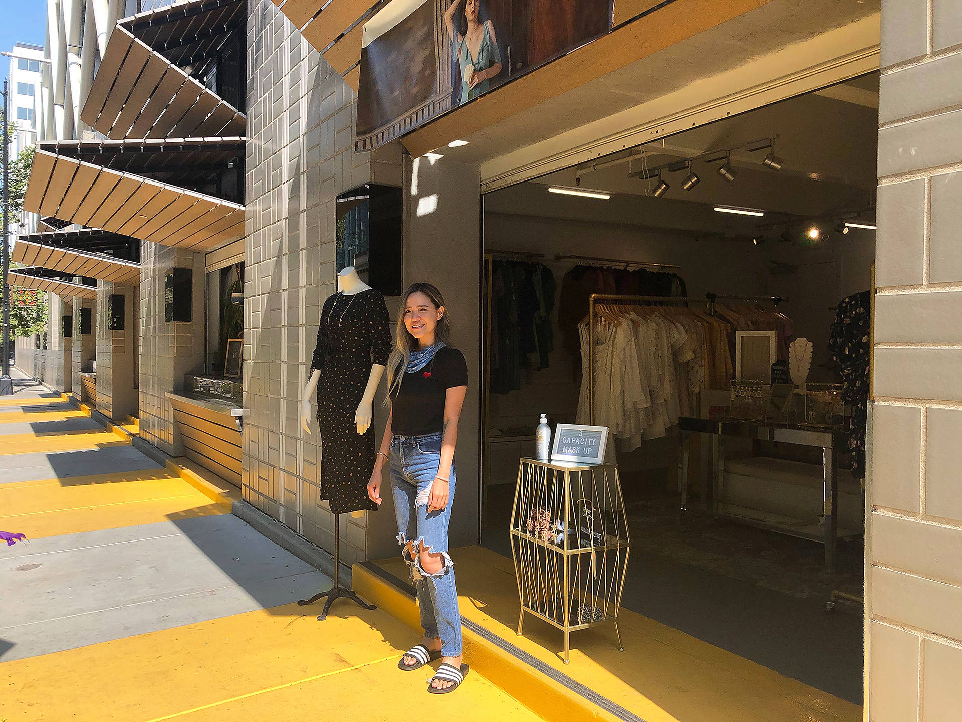 Shopping 桌布 壁紙 — 一位微笑的女士站在一間精品店外，店外有黃色人行道和模特兒公仔，在明亮的日光下。