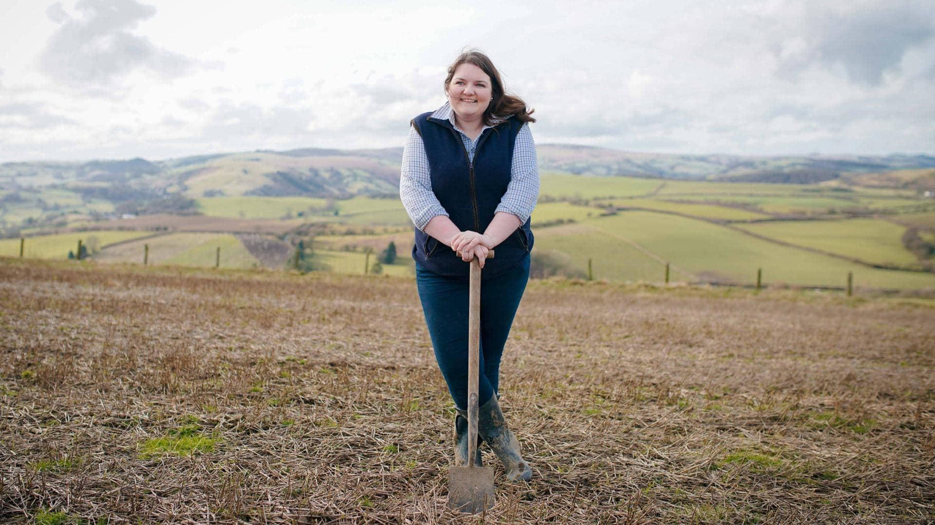 Farmer 桌布 壁紙 — 一位微笑的女士站在乾燥的田野中，手持鏟子，背後是起伏的綠色山丘，天空多雲。