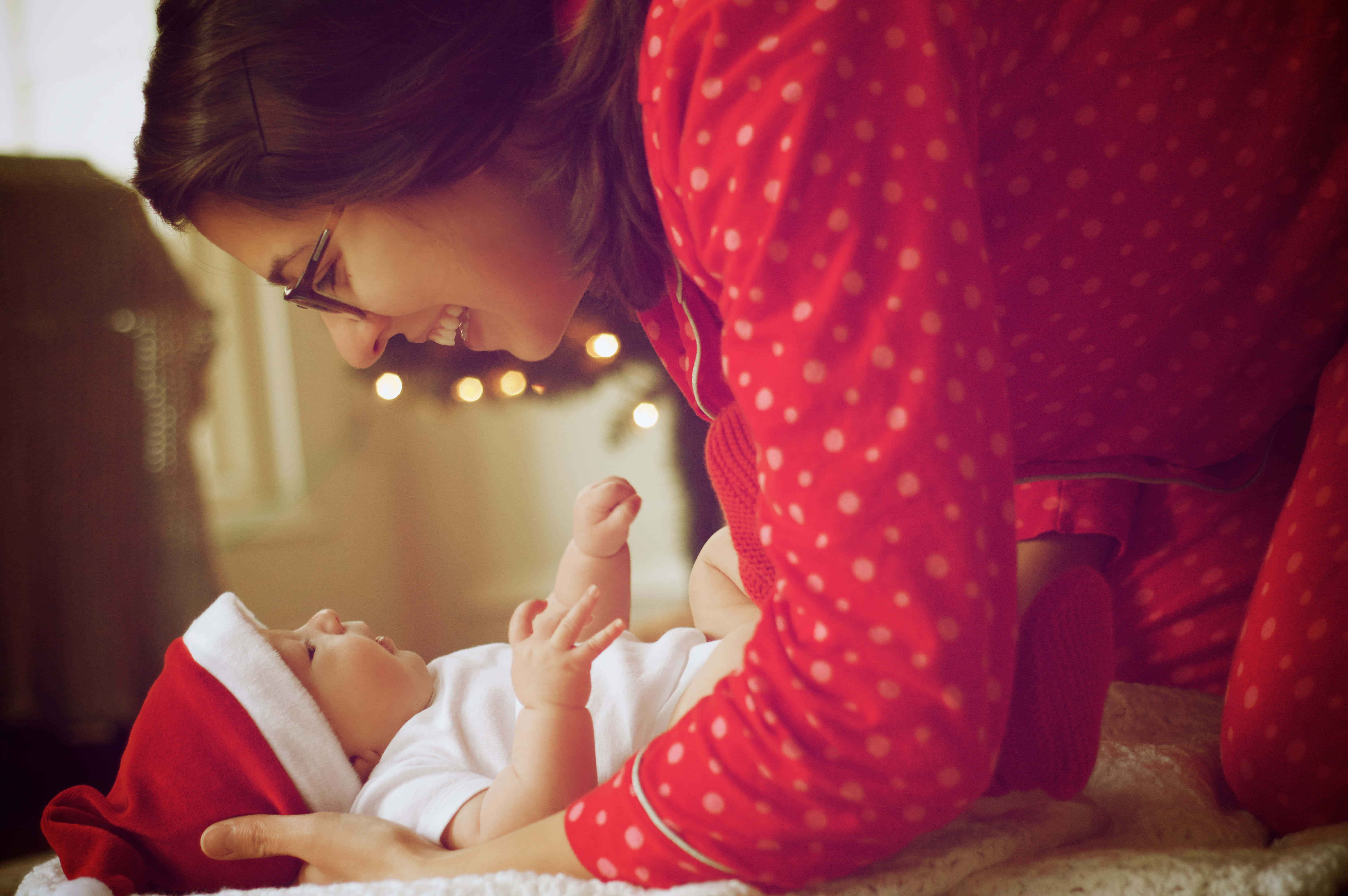 Mom 桌布 壁紙 — 一位微笑的女士身穿紅色圓點睡衣，俯身看著戴著聖誕帽的寶寶，在溫暖舒適的房間裡。