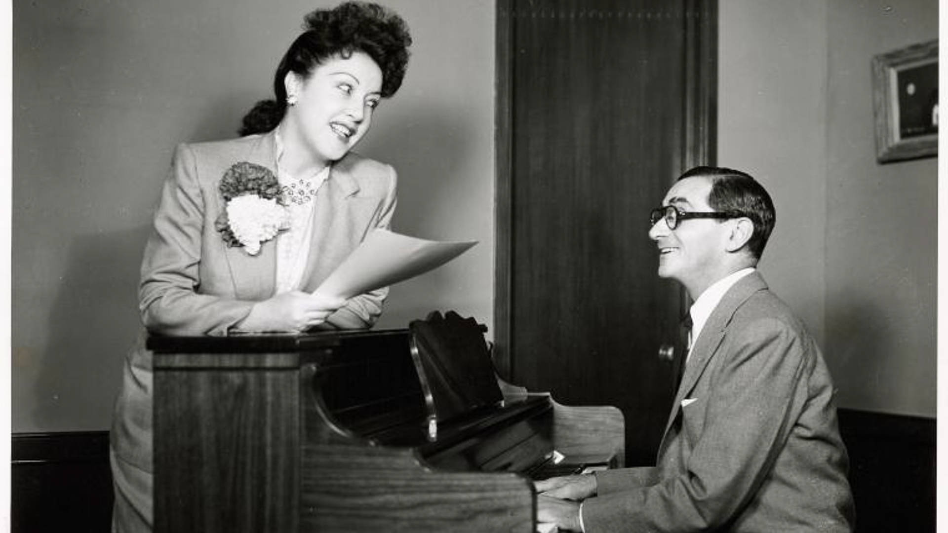 Singing 桌布 壁紙 — 一位微笑的女士身穿西裝站在鋼琴旁，手持樂譜，而一位戴眼鏡的男士正在彈奏並抬頭看著她。