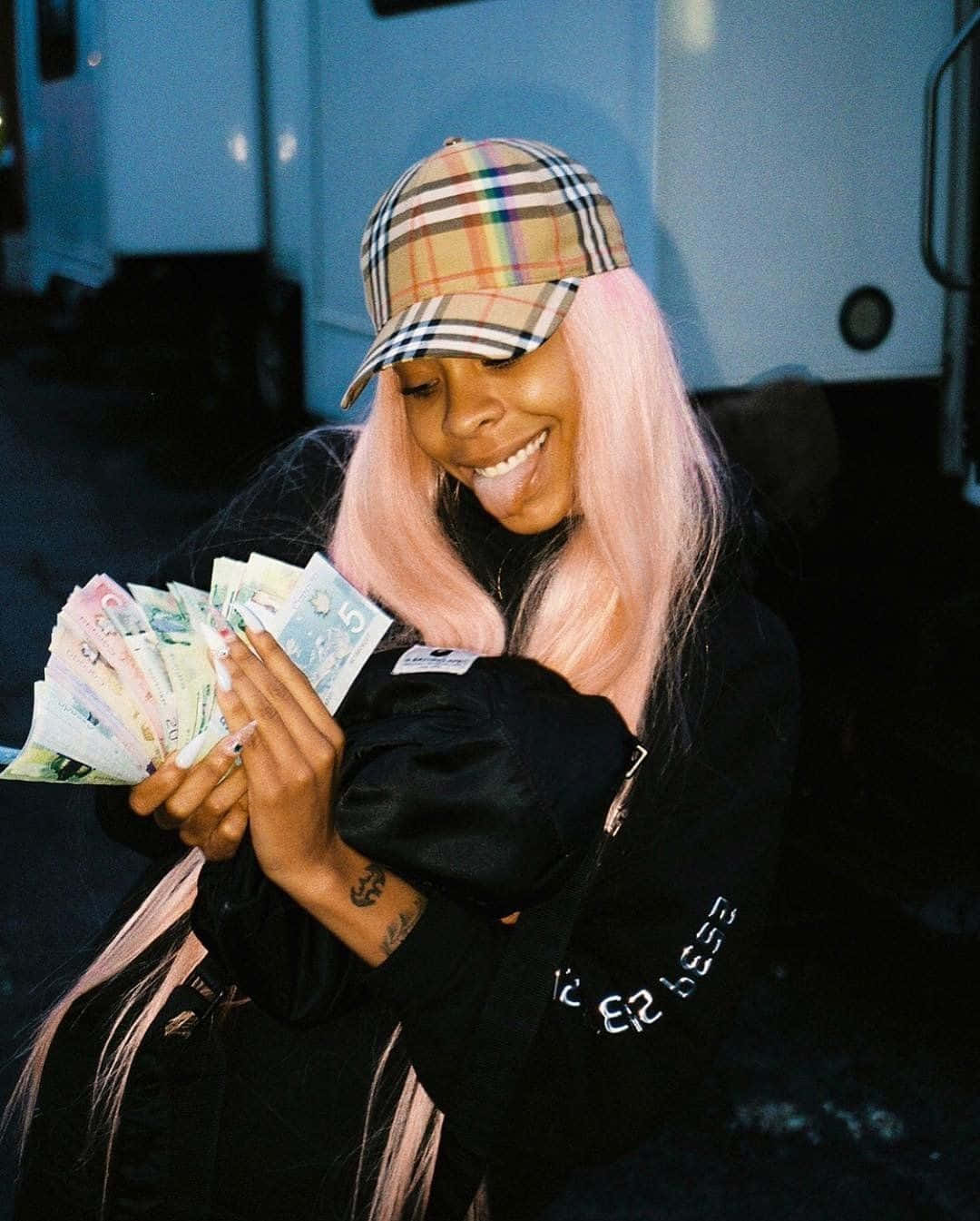 Rico Nasty 桌布 壁紙 — 一位微笑的女子，粉紅色頭髮，戴著格子帽，頑皮地伸出舌頭，同時拿著一把色彩繽紛的鈔票扇。