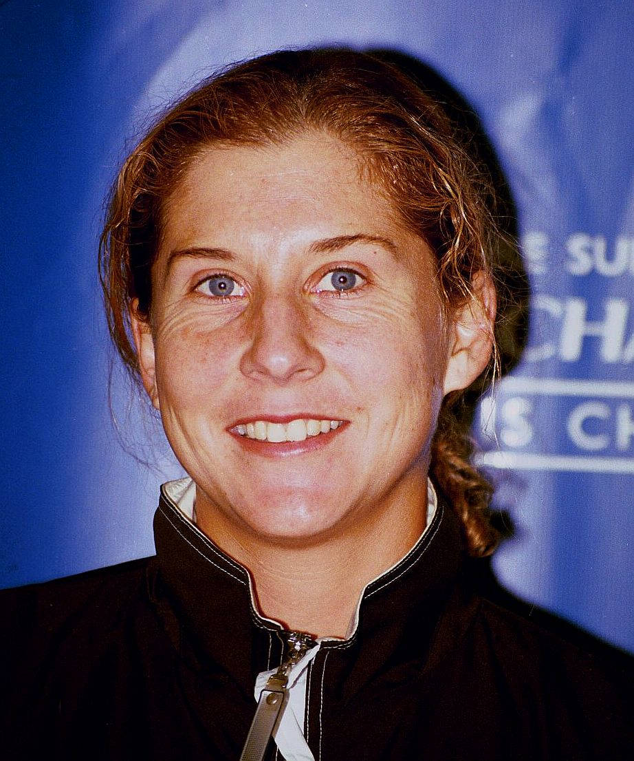 Monica Seles 桌布 壁紙 — 一位微笑的女子，藍眼睛，辮子頭髮，穿著黑色拉鍊外套，背景是藍色活動背景。