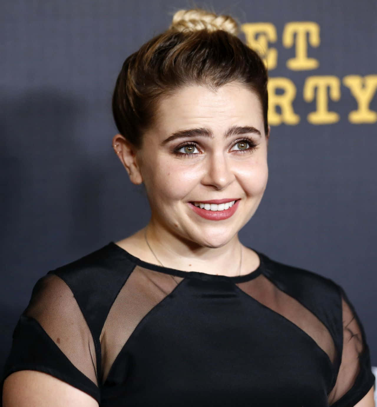 Mae Whitman 桌布 壁紙 — 一位微笑的女子，辮子盤成髮髻，身穿黑色透明袖連衣裙，背景深色並有黃色文字。
