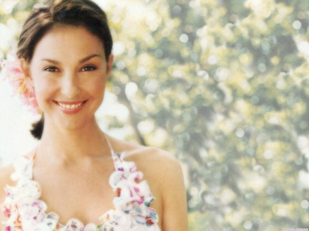 Ashley Judd 桌布 壁紙 — 一位微笑的女子，頭髮上插著一朵粉紅花，穿著花卉圖案上衣，背景是明亮模糊的綠色樹葉。