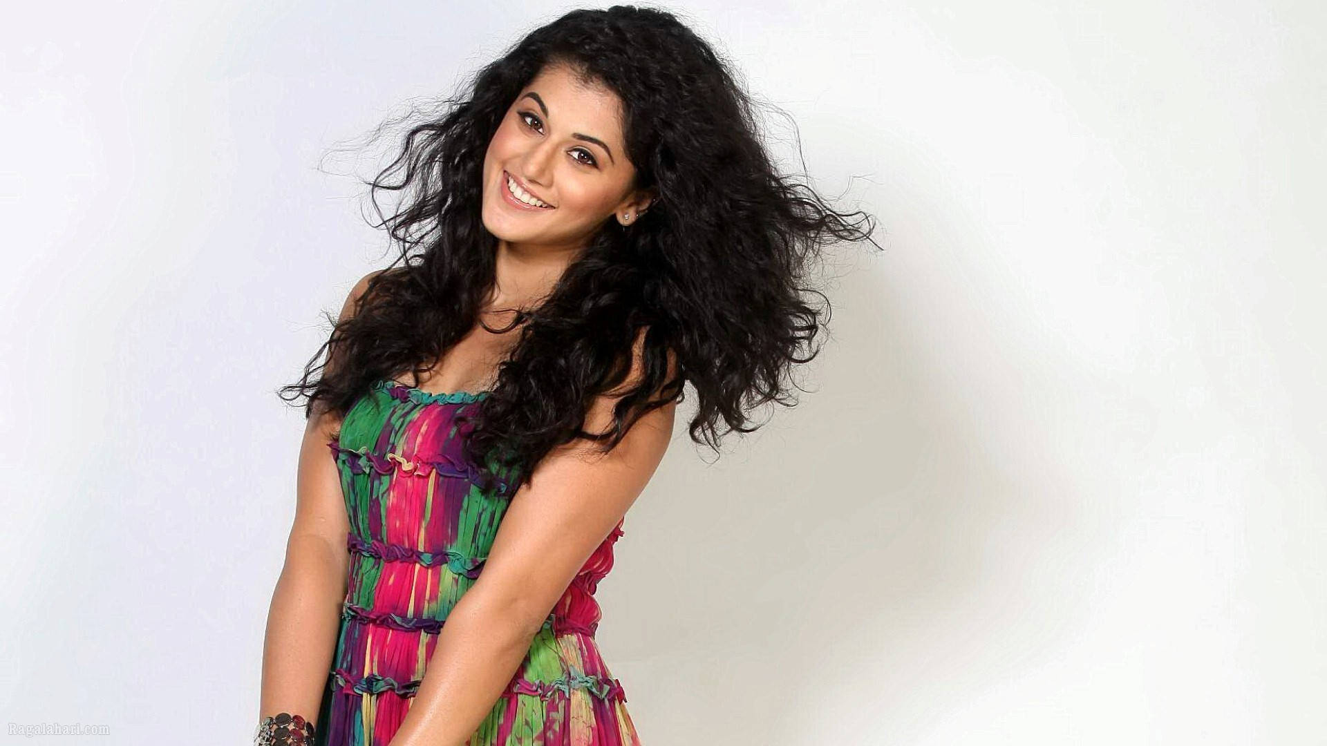 Taapsee Pannu 桌布 壁紙 — 一位微笑的女子，飄逸的深色頭髮，穿著鮮豔多彩的連衣裙，背景是純白色。