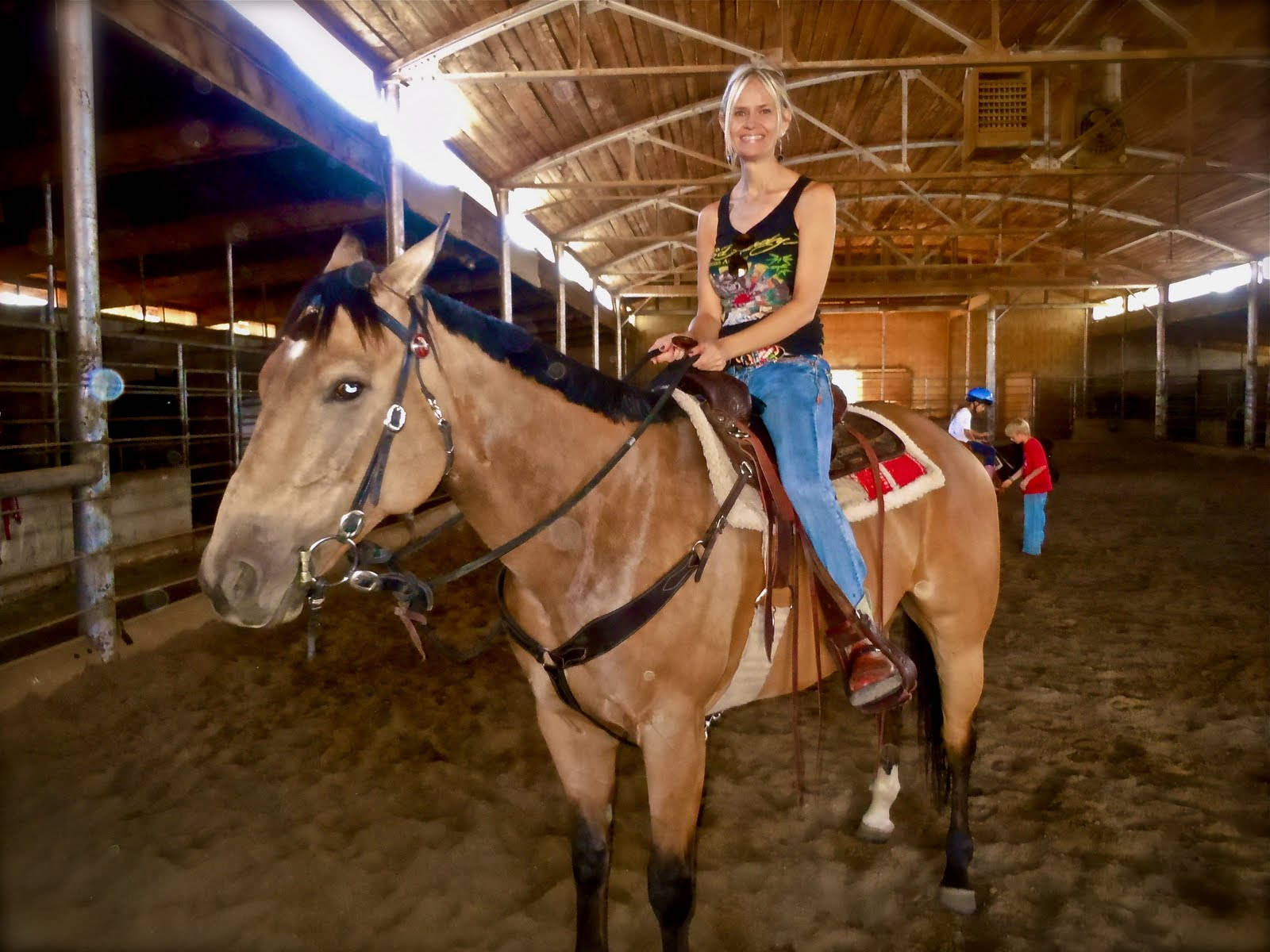 Barrel Racing 桌布 壁紙 — 一位微笑的女子在一個有泥土地板和金屬圍欄的鄉村風格木馬廄裡騎著一匹栗色馬。