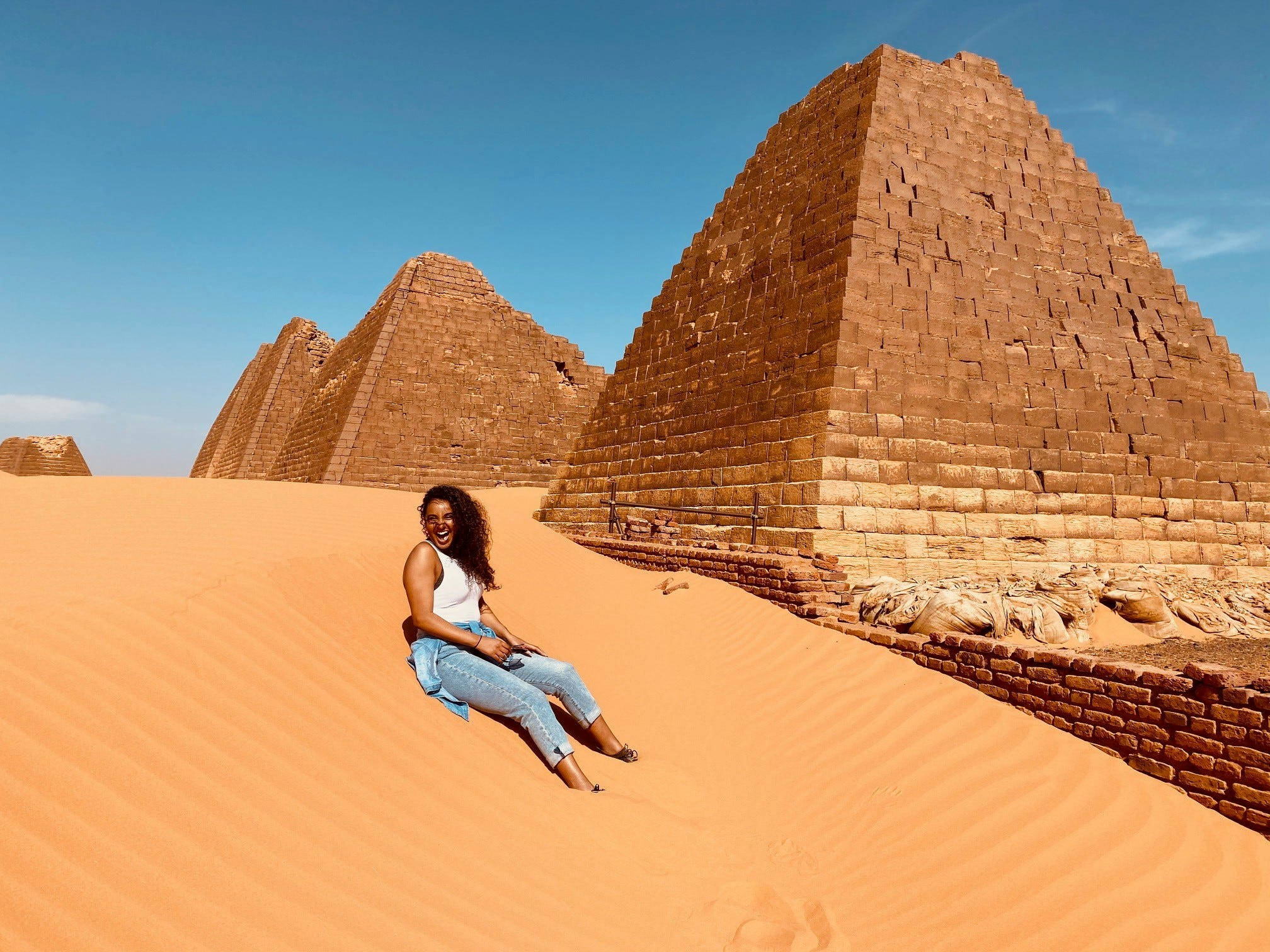 Sudan 桌布 壁紙 — 一位微笑的女子坐在金色沙丘頂上，背後有古老的磚砌金字塔在清澈藍天下聳立。