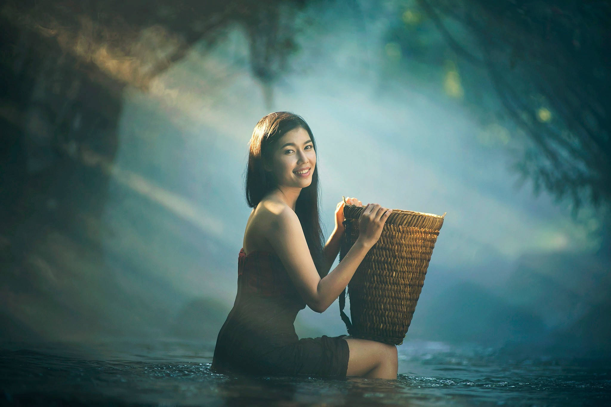 美女 桌布 壁紙 — 一位微笑的女子坐在霧氣瀰漫的水中，手持一個編織籃，陽光穿過她身後的森林樹冠照射下來。