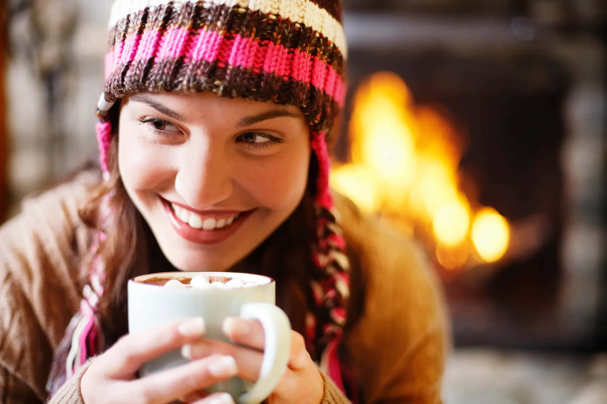 Hot Chocolate 桌布 壁紙 — 一位微笑的女子戴著彩色針織帽，在溫暖發光的壁爐旁拿著一杯熱可可。
