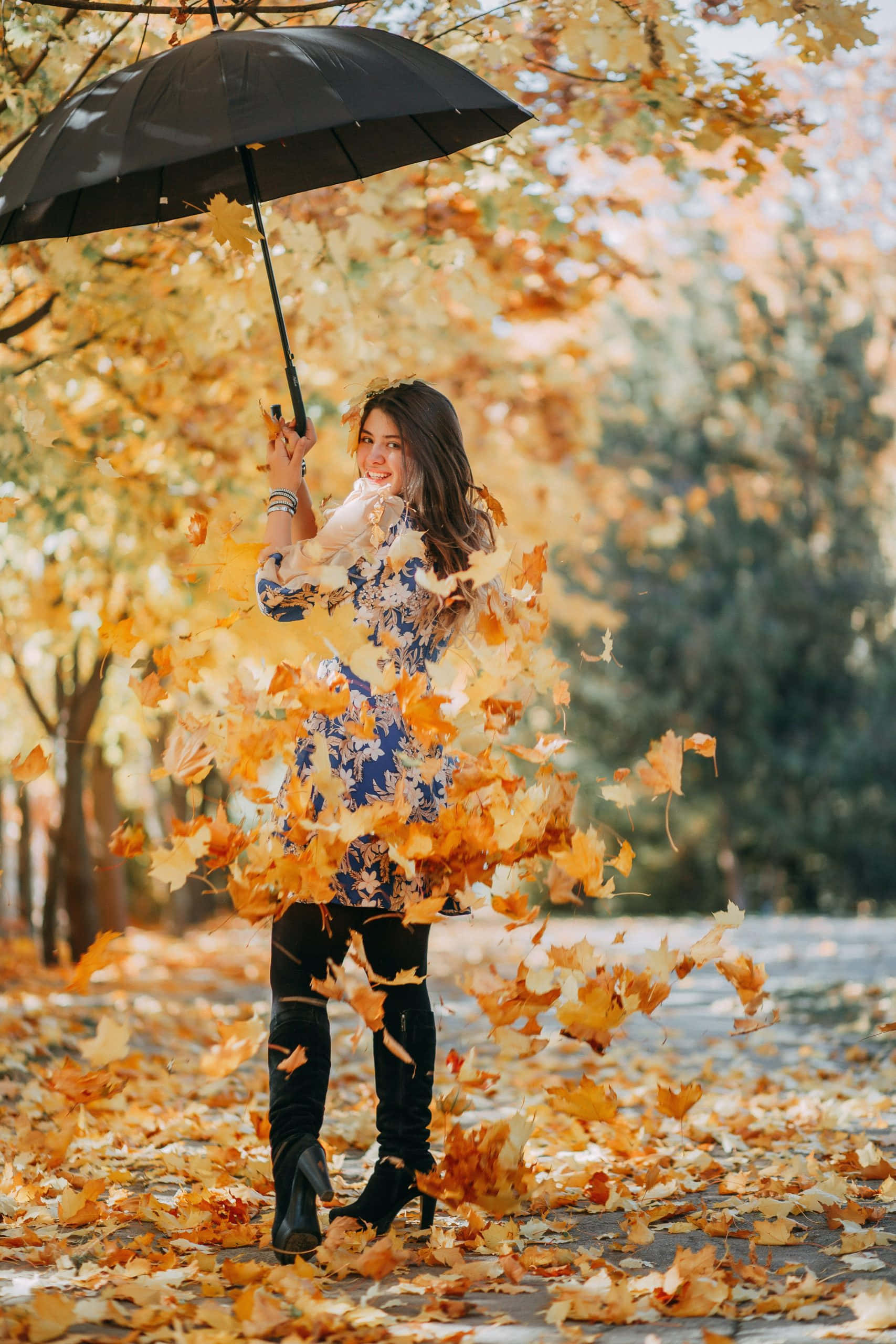 Fall Photography 桌布 壁紙 — 一位微笑的女子手持黑色雨傘，金色秋葉在她周圍的公園中旋轉飄落。