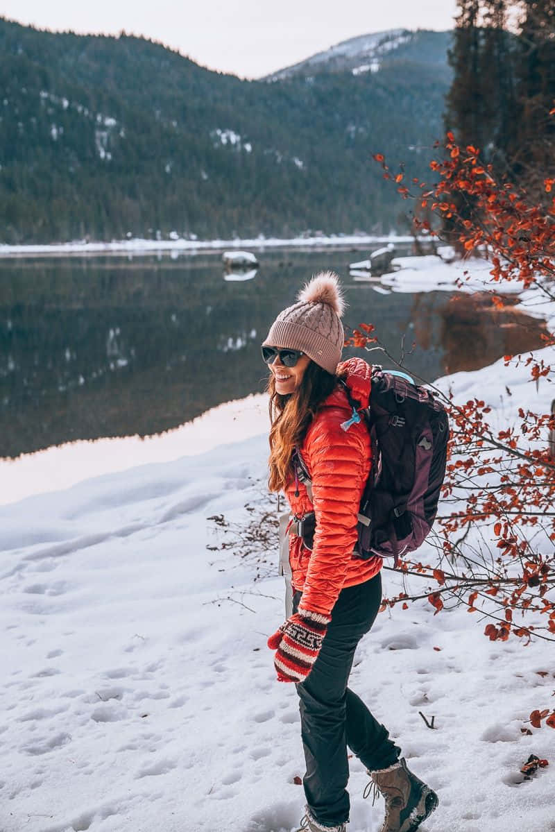 Winter Hiking 桌布 壁紙 — 一位微笑的女子穿著亮橙色外套和粉紅色毛帽，在雪覆蓋的森林中行走，旁邊是平靜的湖泊，背景是白雪覆蓋的山脈。