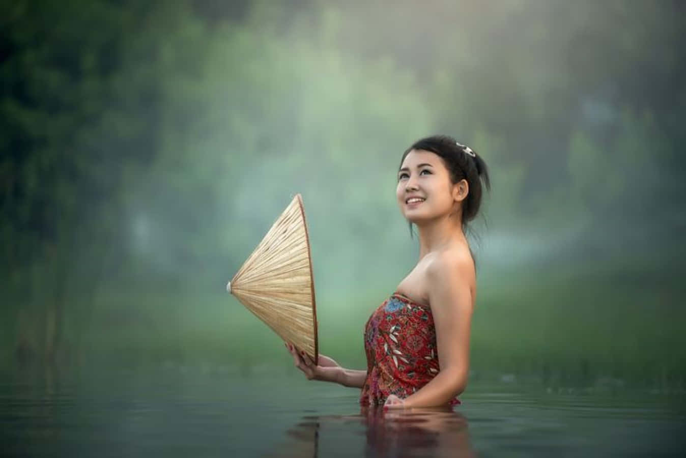美女 桌布 壁紙 — 一位微笑的女子穿著圖案連衣裙，站在霧氣瀰漫的水中，水深及腰，手持一頂圓錐形帽子，身後是茂密的綠植。