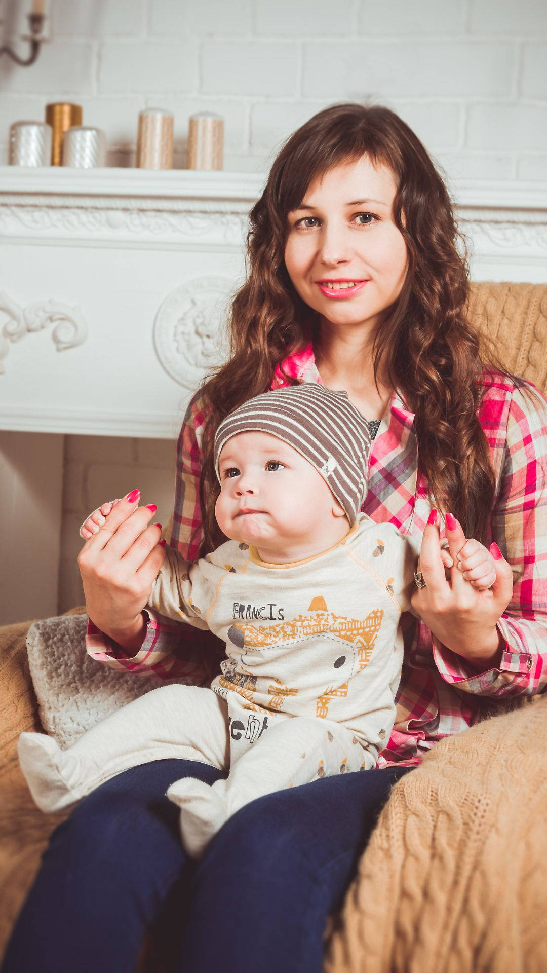 Mom 桌布 壁紙 — 一位微笑的女子穿著格紋襯衫，在白色壁爐附近舒適的針織沙發上抱著一個戴著條紋帽子的嬰兒。