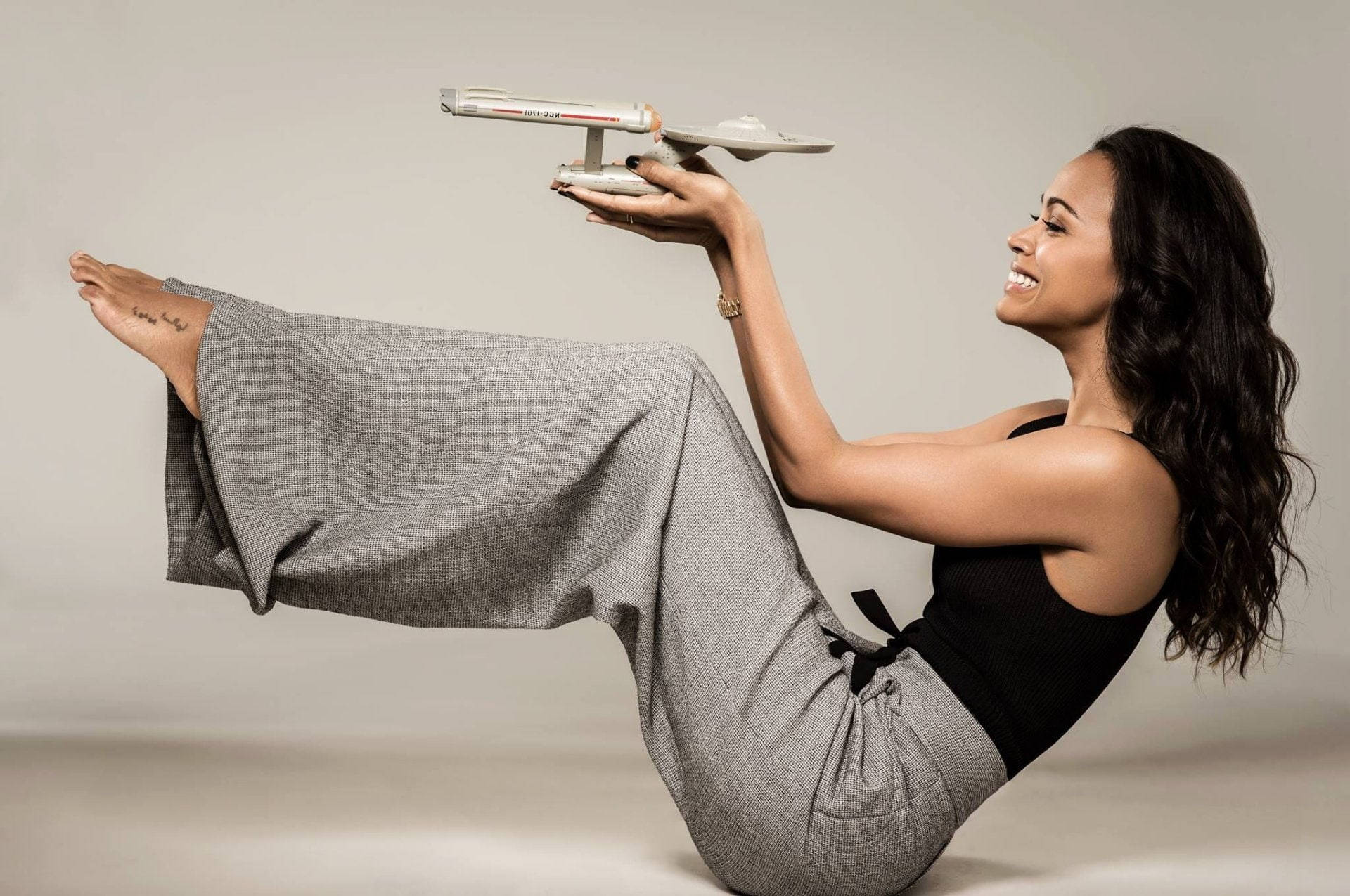Zoe Saldana 桌布 壁紙 — 一位微笑的女子穿著灰色褲子，坐在中性的攝影棚地板上，手上平衡著星際爭霸戰的飛船模型。