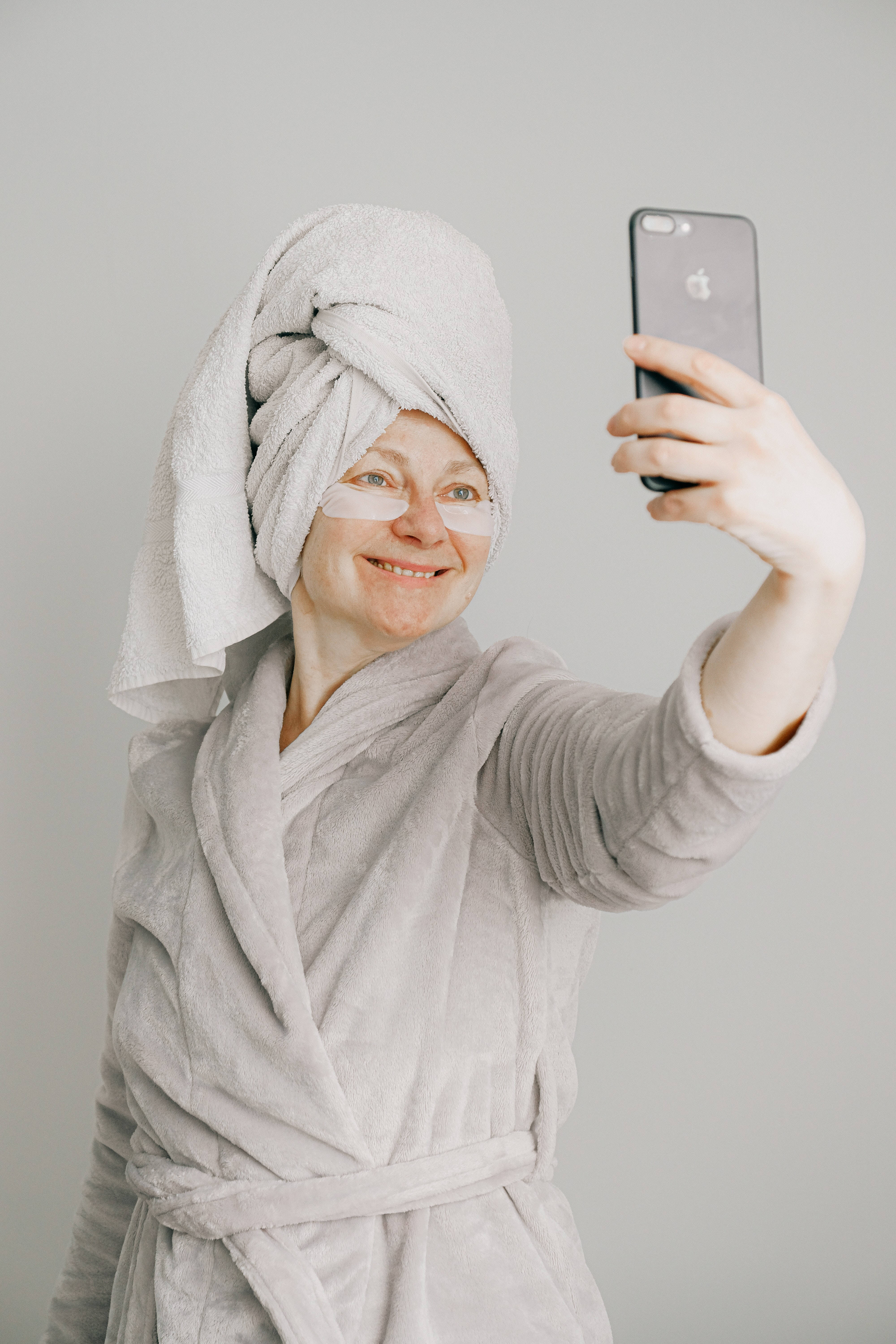 iPhone 桌布 壁紙 — 一位微笑的女子穿著灰色長袍，頭上裹著毛巾，貼著眼膜，在純灰色背景前用手機自拍。