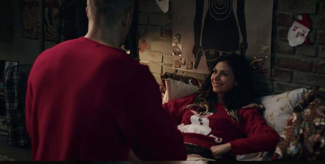 Deadpool And Vanessa 桌布 壁紙 — 一位微笑的女子穿著紅色雪人毛衣坐在床上，面對一位穿著紅色襯衫的男子，房間舒適昏暗，有磚牆和節日裝飾。