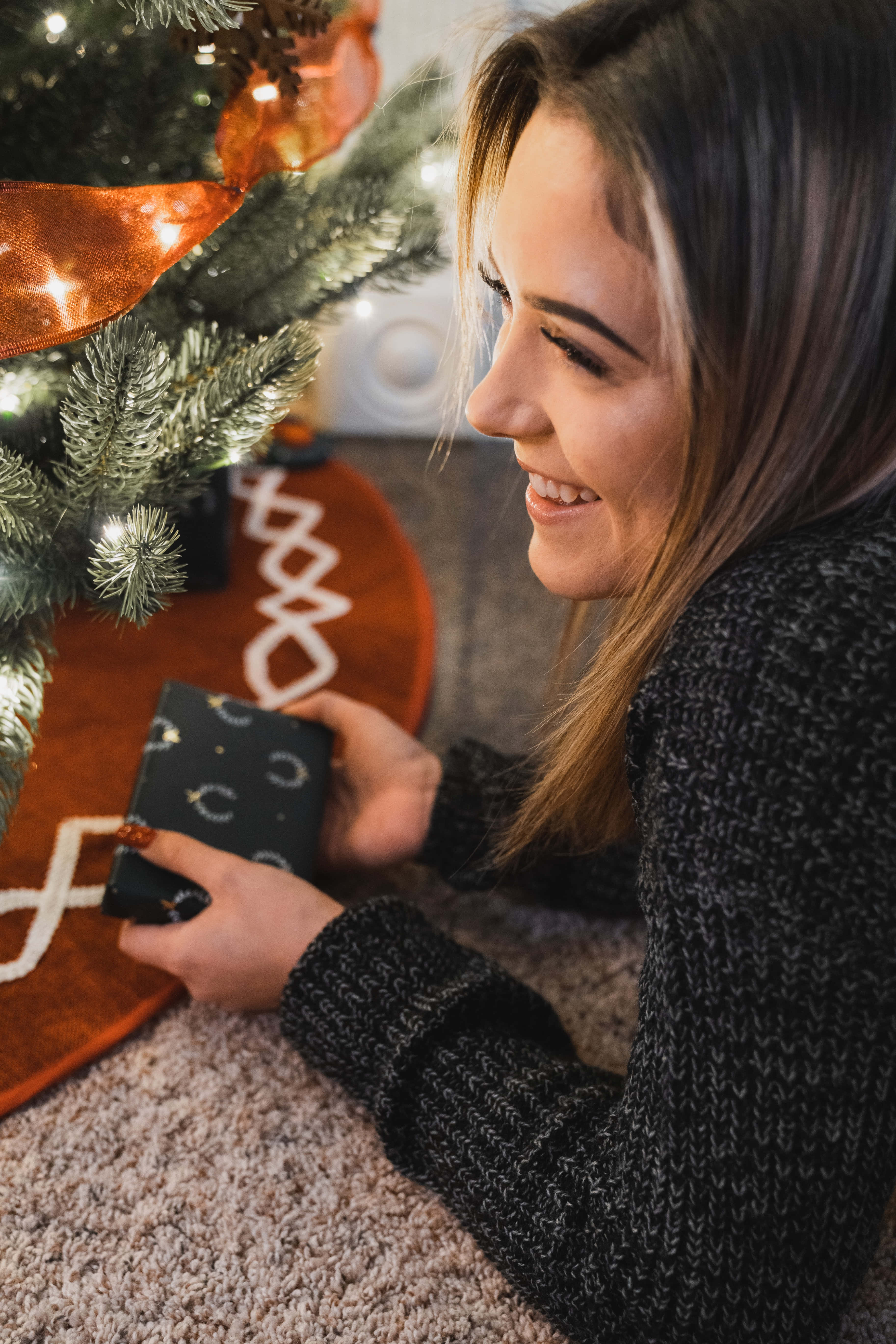 Christmas Scene 桌布 壁紙 — 一位微笑的女子穿著舒適的毛衣，在裝飾著溫暖燈光的聖誕樹旁拿著一份小禮物。