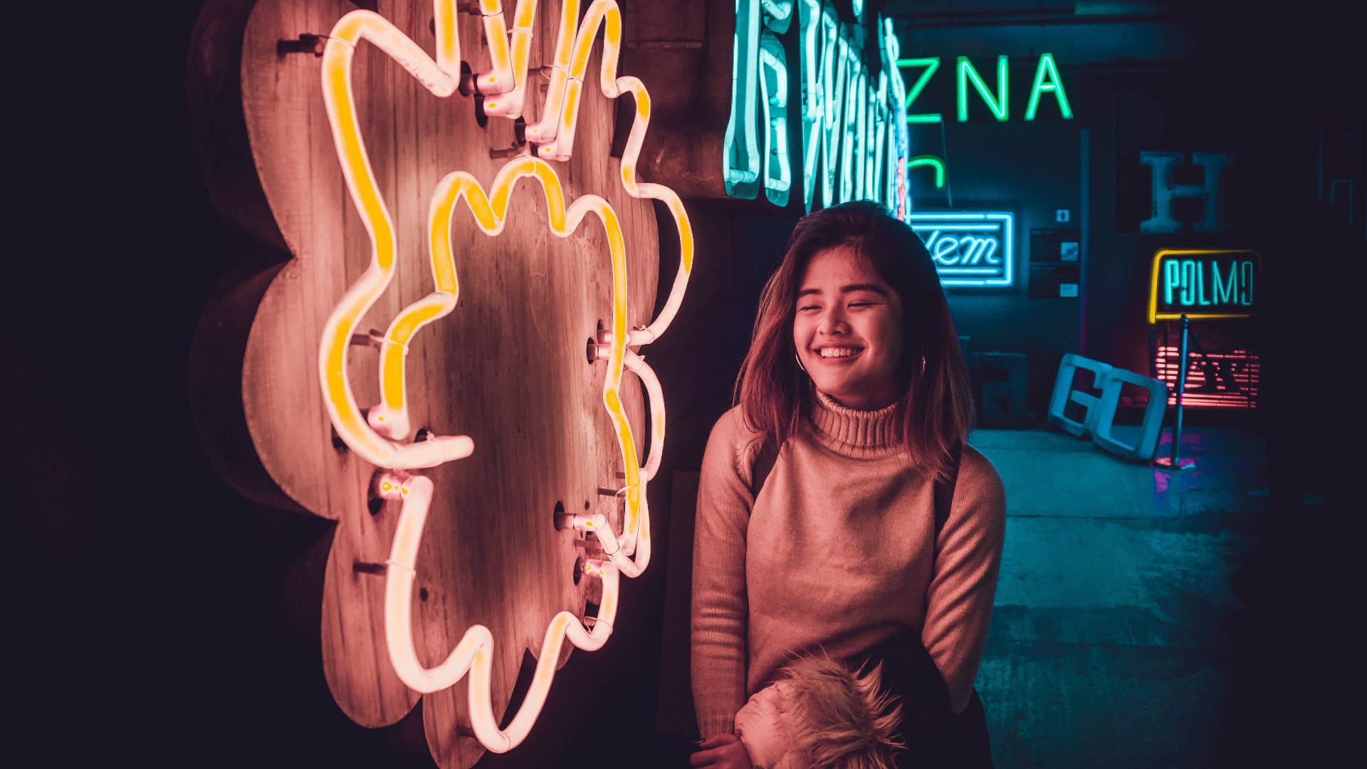 Neon Light 桌布 壁紙 — 一位微笑的女子穿著舒適的毛衣，站在一條昏暗的城市小巷中，旁邊是一朵發光的黃色霓虹花，周圍有色彩繽紛的霓虹招牌。