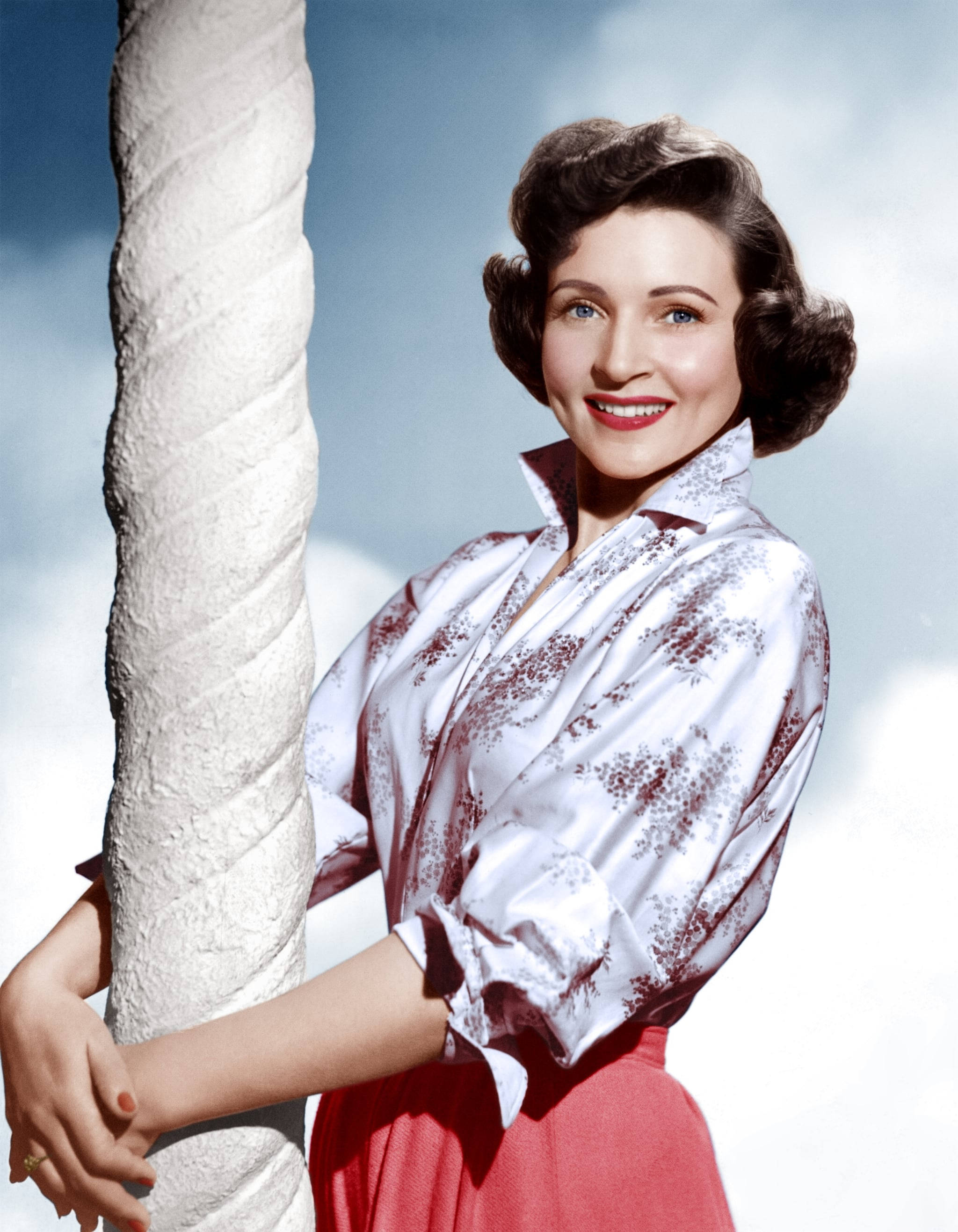 Betty White 桌布 壁紙 — 一位微笑的女子穿著花卉圖案上衣和紅色裙子，靠在一根扭曲的白色柱子上，背景是明亮的天空。