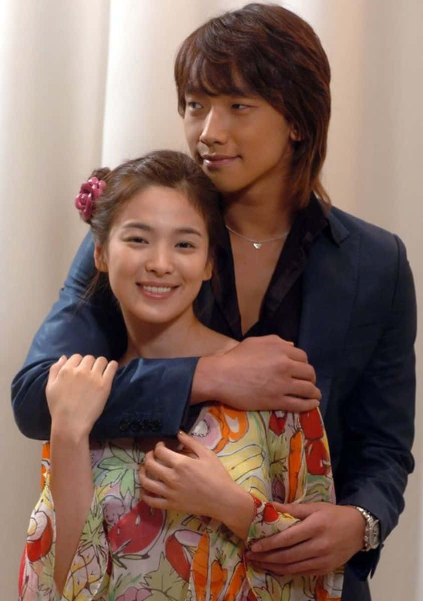 Beautiful Couple 桌布 壁紙 — 一位微笑的女子穿著花卉連衣裙，被一位穿著深色西裝的男子在柔和的米色窗簾背景下溫暖地擁抱。