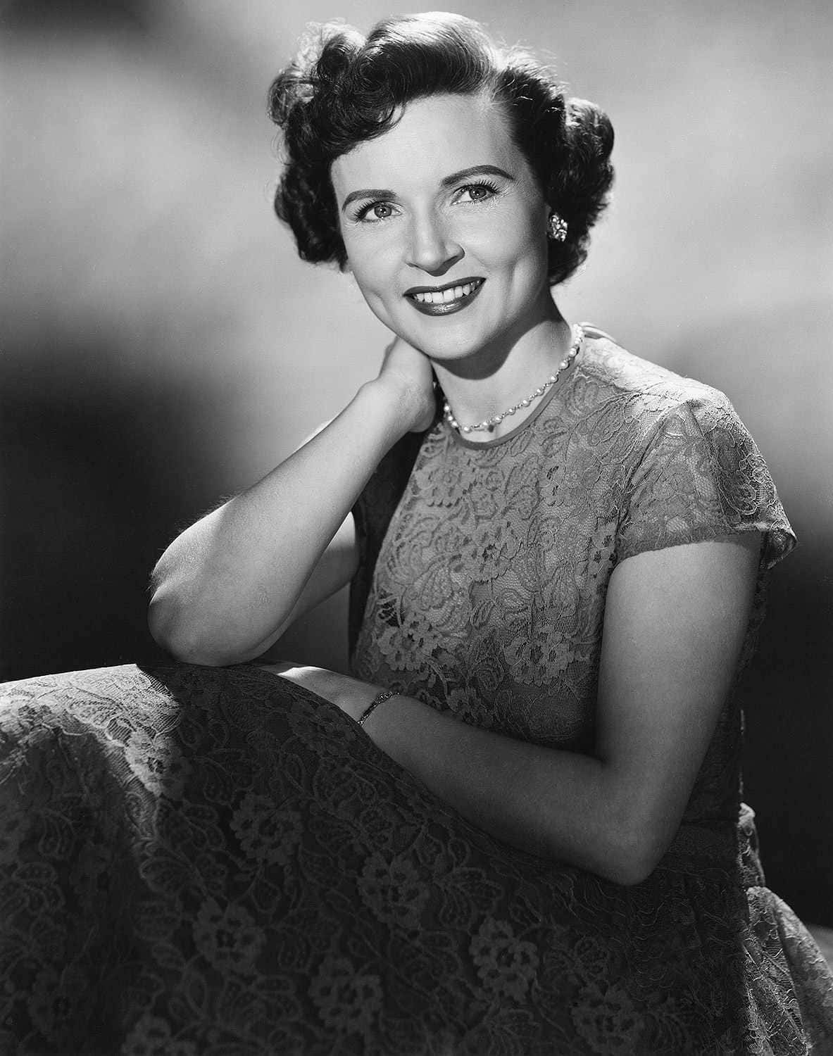 Betty White 桌布 壁紙 — 一位微笑的女子穿著蕾絲裙，一手放在頸上，在柔和模糊的攝影棚背景下優雅地擺姿勢。