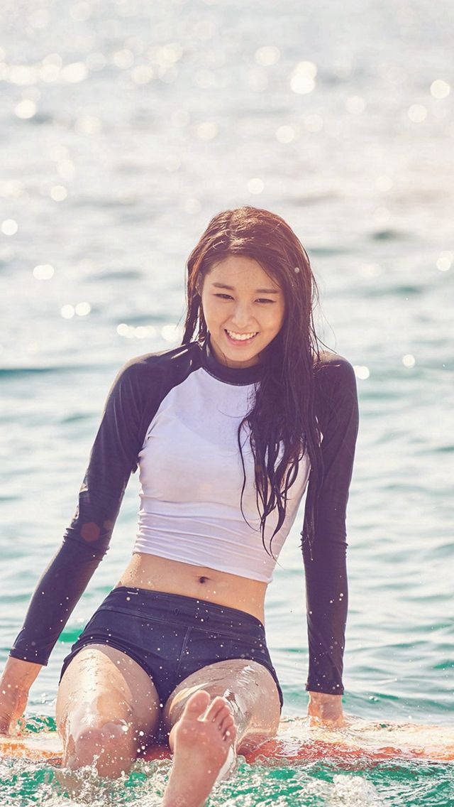 美女 桌布 壁紙 — 一位微笑的女子穿著雙色防曬衣，坐在陽光下閃閃發光的海水中的衝浪板上。