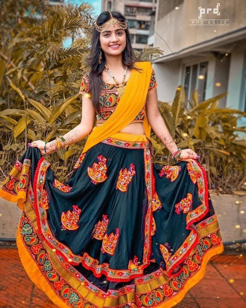 Nisha 桌布 壁紙 — 一位微笑的女子穿著鮮豔的傳統印度 lehenga，帶有金色和橙色刺繡，在戶外鬱鬱蔥蔥的綠植中擺姿勢。