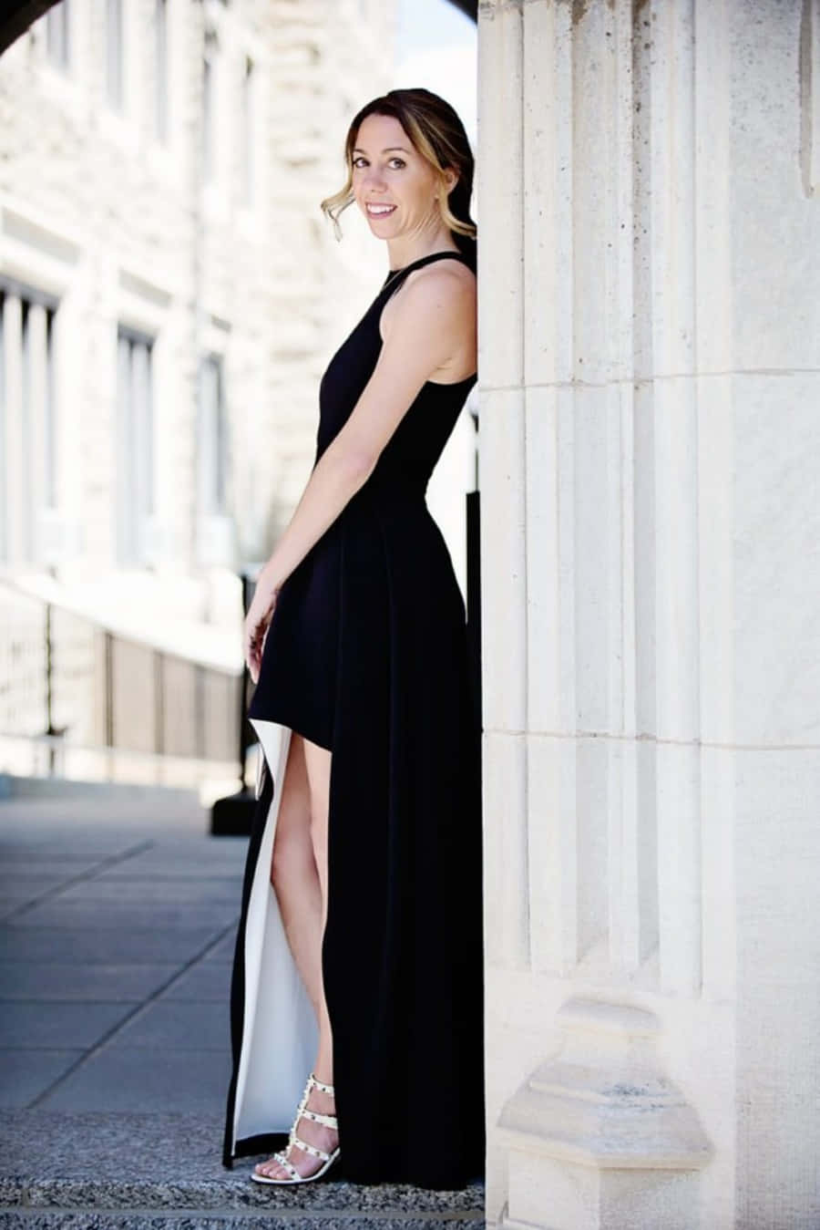 Black Tie Dresses 桌布 壁紙 — 一位微笑的女子穿著黑色高衩連衣裙，在陽光明媚的城市人行道上靠著石柱。