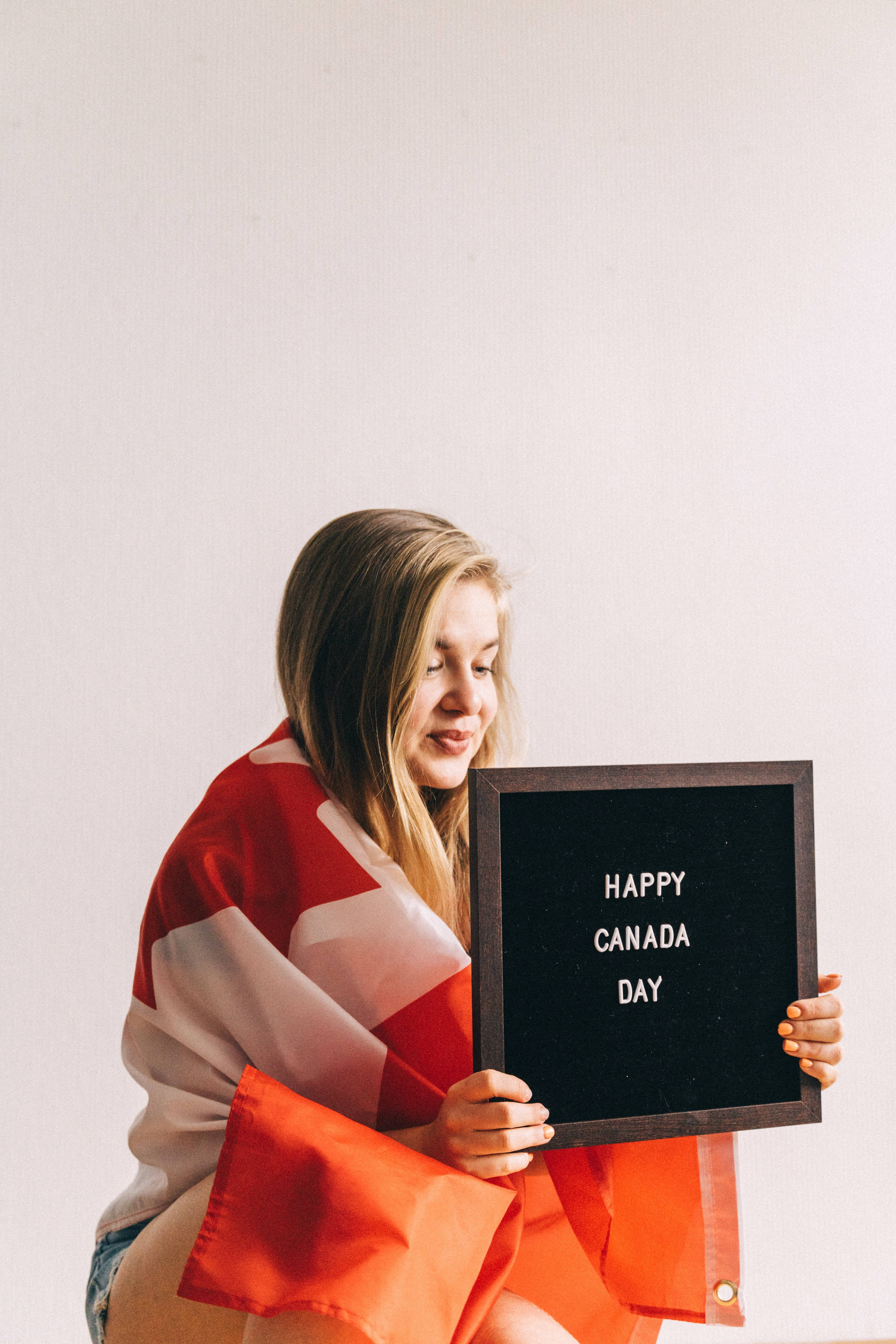 Greetings 桌布 壁紙 — 一位微笑的女子裹著加拿大國旗，手持寫有「HAPPY CANADA DAY」的標語牌，背景為純白色。